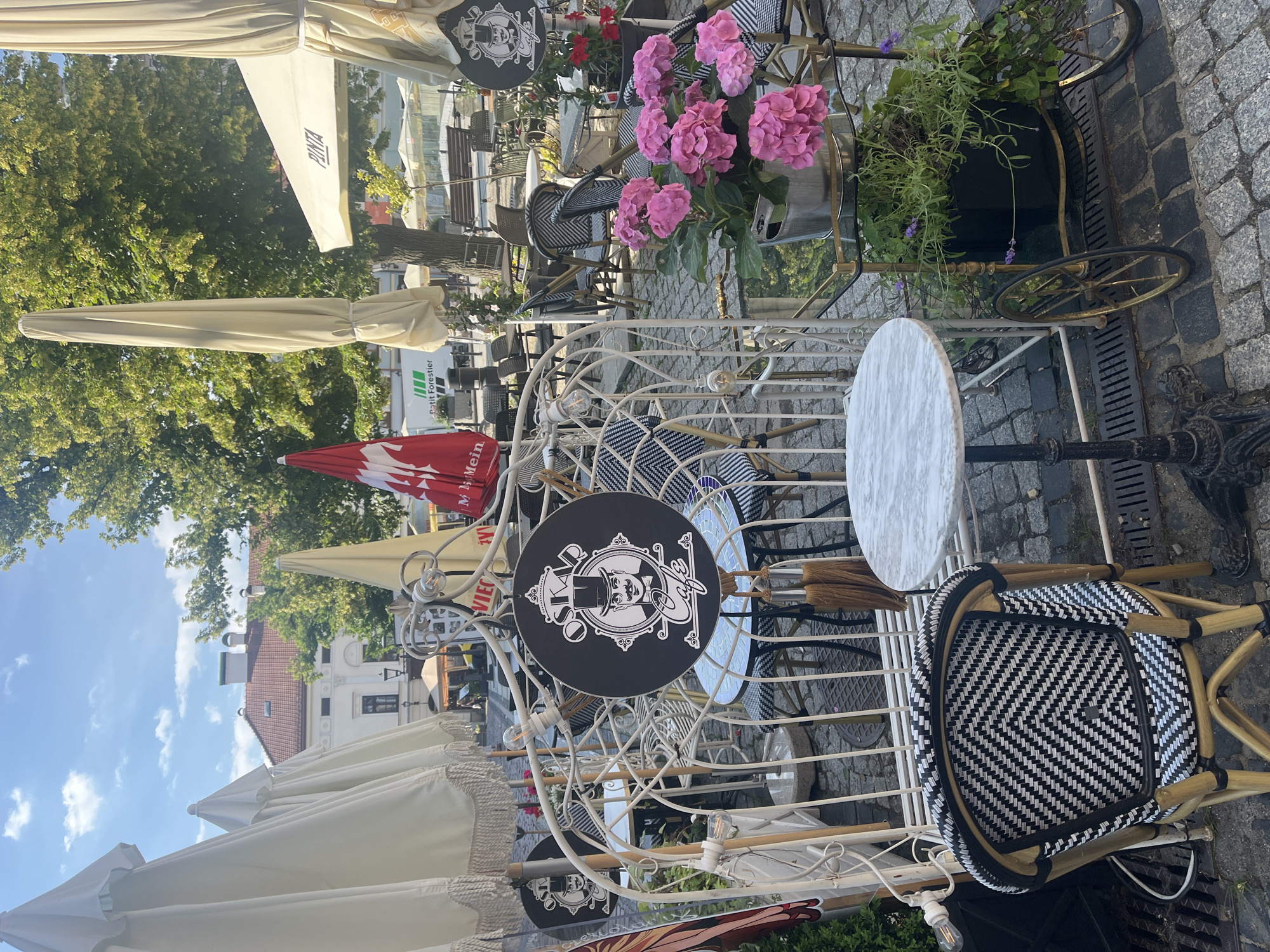 An outdoor seating area for a cafe is shown on a cobblestone street. Several white and beige umbrellas are set up, along with tables and chairs. A decorative white metal structure, resembling a gazebo or archway, is in the foreground. A sign for "Osk" cafe with a stylized drawing of a man in a top hat hangs from this structure. Several tables with black and white checkered chairs are visible. A potted pink hydrangea and other greenery are placed on a rolling cart with wheels. In the background, more outdoor seating, trees, and buildings are visible. The sky is blue with some clouds.