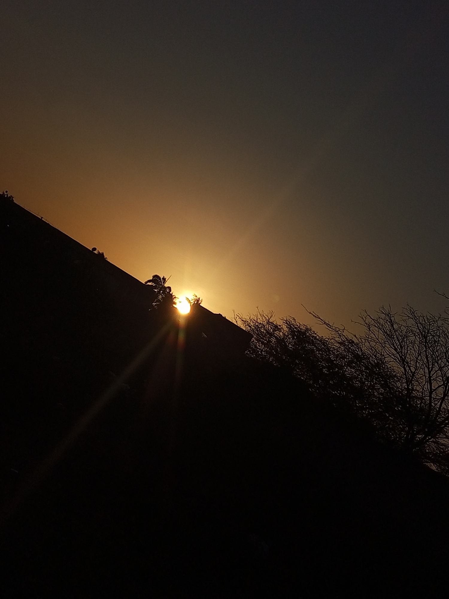 The image captures a sunset scene in Juan Griego, Venezuela. The sun, low on the horizon, casts a bright light through a dark silhouette of trees and possibly a hill. The sky transitions from a dark grey-blue to a warm golden hue near the horizon. A few rays of sunlight are visible, streaking down from the sun. The foreground is predominantly dark, obscuring details.