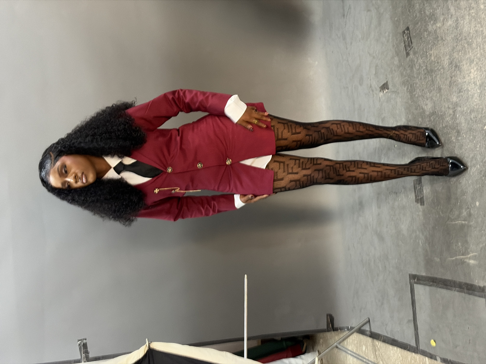A woman is depicted standing in what appears to be a studio setting in the city of Gura Top, Nigeria. She stands facing forward, with her head turned to her right (viewer's left), looking away from the camera. Her complexion is dark, and she has long, dark, curly hair styled with a defined side part. She is wearing a burgundy blazer-style dress featuring a white collar and cuffs, a black tie, and three gold buttons vertically aligned on the front. A gold chain ornament with a cross-like element is pinned to the left lapel of the blazer. She wears sheer black tights with a repeating 'F' pattern, indicative of the Fendi brand. Her feet are clad in black patent leather pointed-toe high-heeled shoes. Rings are visible on the fingers of her right hand.
The background consists of a plain, solid grey wall. The floor is a textured grey surface, possibly concrete, with visible marks and dark lines forming rectangular shapes, suggesting floor tiles or sections. In the bottom left corner, a small yellow object rests on the floor. To the left of the woman, studio equipment is partially visible, including a light stand with a softbox reflector and a thin horizontal metal rod extending towards the center of the frame. The overall lighting appears even, suggesting controlled studio conditions.