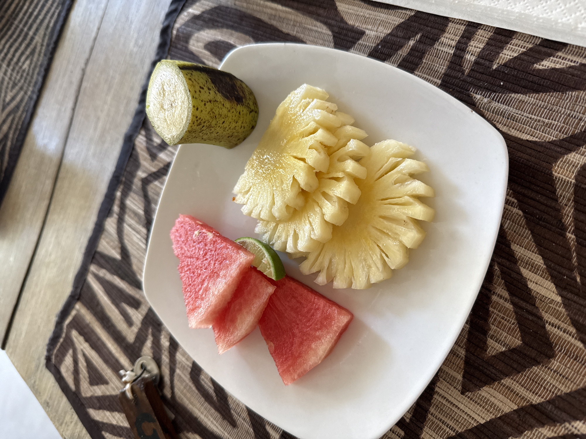 A white plate with fresh fruit sits on a patterned surface. The fruit includes sliced pineapple, watermelon, and a section of a banana. A thin slice of lime rests between the watermelon pieces. The location is Unknown, Indonesia.