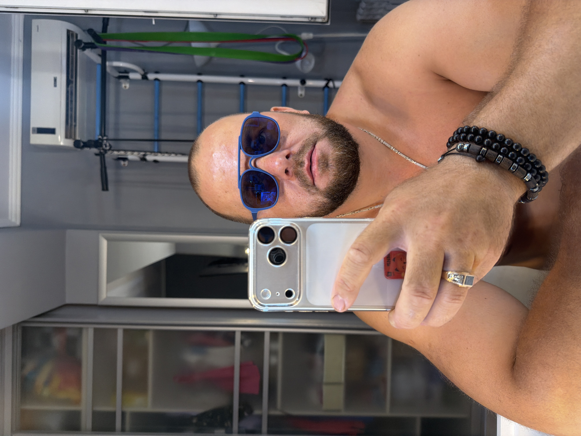 This is a selfie taken by a man in what appears to be a home gym or workout space, likely in Sochi, Russia. The man is shirtless and wearing blue aviator sunglasses and several beaded bracelets on his left wrist. He also wears a ring on his left index finger and a silver chain around his neck. The background shows workout equipment, including resistance bands, and a closet with shelves containing various items. The lighting suggests it could be daytime. The man's expression is neutral. The image is oriented sideways, and the camera is held by the man to capture the reflection.
