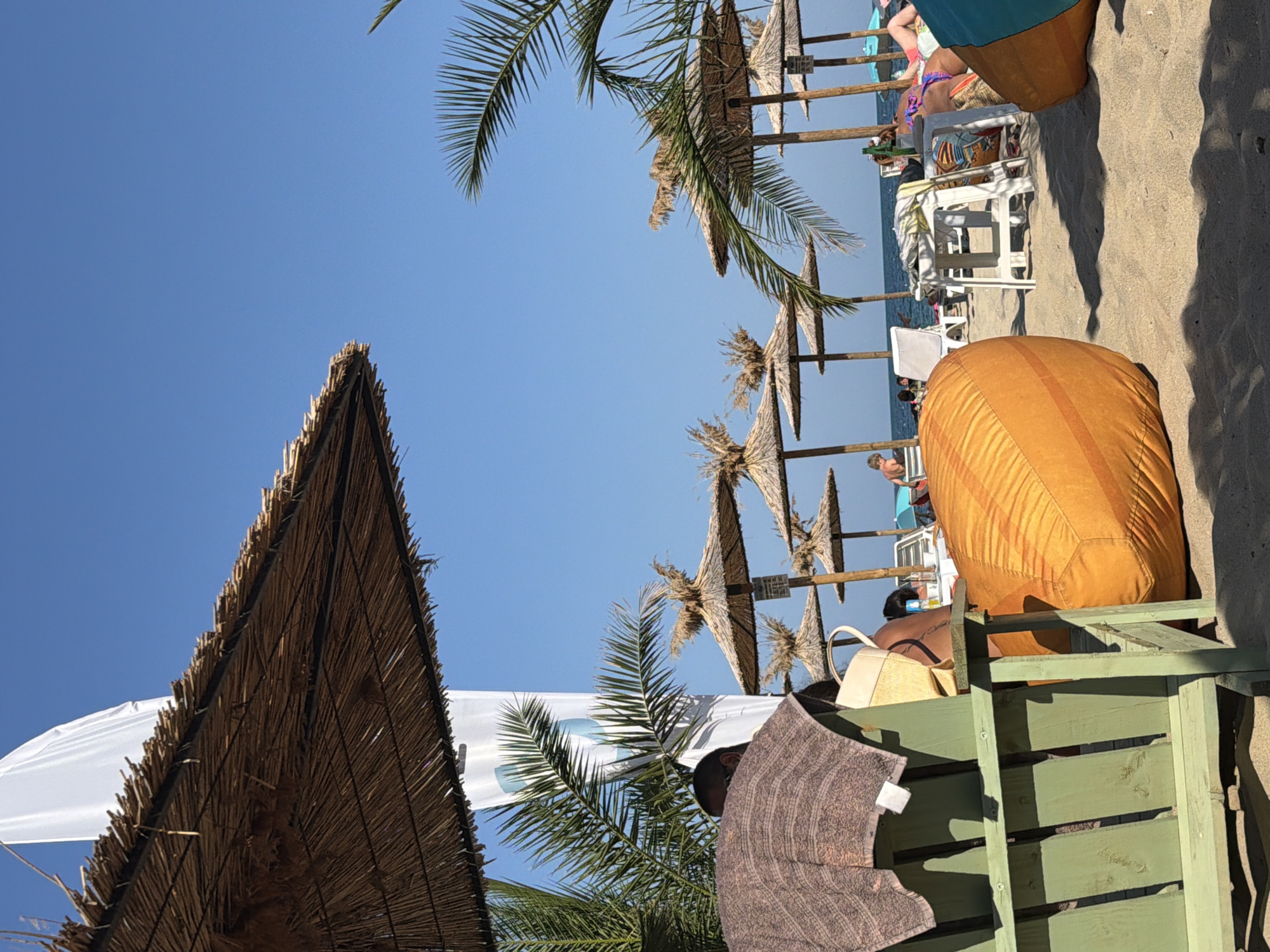 This image captures a sunny day at a beach, likely in Varna, Bulgaria, given the context. In the foreground, a green wooden chair is partially visible, draped with a brown towel. Beside it rests a large, orange beanbag chair. Further into the scene, numerous thatched umbrellas provide shade over the sandy beach. Several beach chairs and lounge chairs are arranged beneath these umbrellas, some occupied by people relaxing. In the background, the calm sea meets the clear blue sky. Palm trees add a tropical feel to the environment. The overall atmosphere is one of leisure and a typical summer day at the seaside.