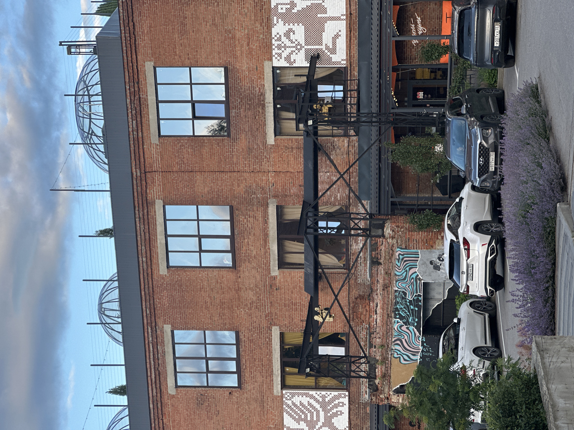 The image depicts the exterior of a brick building with large, multi-paned windows. The building has a modern architectural element on its roof, featuring geodesic dome structures. In front of the building, there is a parking area with several cars, including white SUVs and a dark gray SUV. There is also a patch of purple flowers in the foreground. The overall scene suggests an urban or suburban setting, possibly a commercial or residential complex. The graffiti on the lower part of the building and the outdoor seating area suggest a restaurant or entertainment venue. The image does not contain any specific landmarks or clues that would allow for precise identification of the city as Vladimir, Russia.