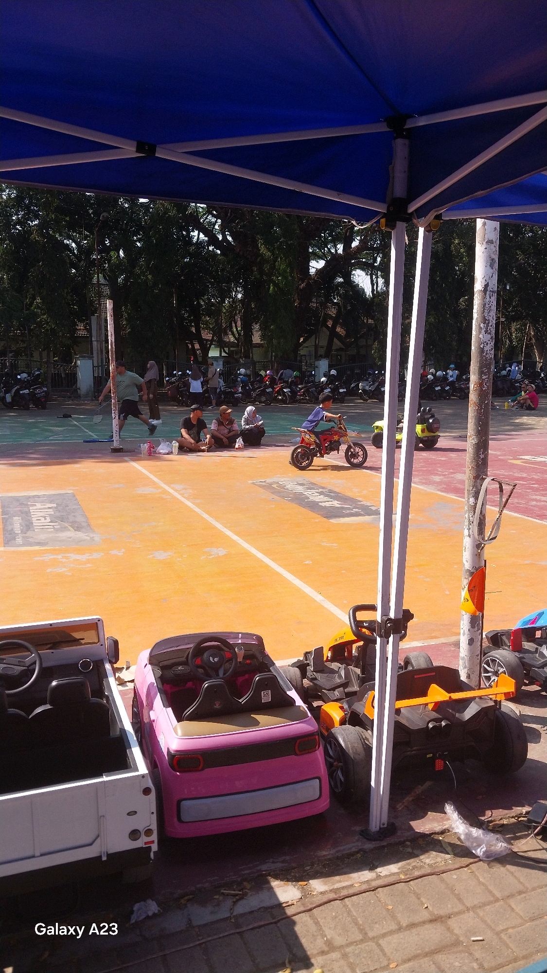The image captures an outdoor scene likely in the city of Malang, Indonesia, under a blue canopy. In the foreground, several parked electric toy cars are visible: a white open-top vehicle, a pink convertible, and a black and orange sports car. Behind these, a basketball court with an orange surface is laid out, marked with white lines.
On the court, a young boy is riding a red and white electric motorcycle, while another yellow electric vehicle is parked nearby. Several people are seated on the court, watching the activities, and a man appears to be playing badminton in the background. Beyond the court, numerous motorcycles are parked, suggesting a public gathering or event. Trees line the perimeter of the area, and a white pole with some attachments stands prominently in the midground. The scene is illuminated by sunlight, casting shadows on the ground. A watermark "Galaxy A23" is visible in the bottom left corner.