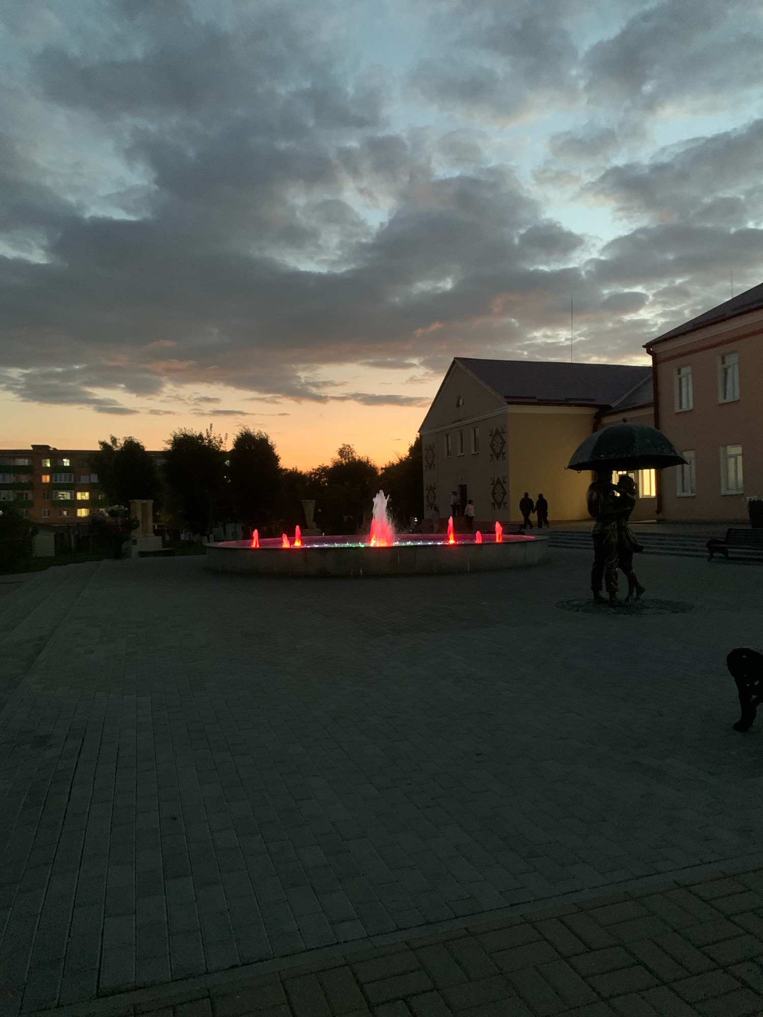 At dusk in Kapyĺ, Belarus, a town square features a circular fountain with colorful red and green lights illuminating the water jets. To the right of the fountain, a bronze sculpture depicts a couple embracing under an umbrella. In the background, two- and three-story buildings with illuminated windows are visible. The sky is filled with dramatic clouds, with the warm hues of sunset peeking through the horizon. The ground is paved with interlocking bricks, and a few people are scattered throughout the scene, with two figures walking hand-in-hand near the fountain and another couple strolling towards the buildings.
