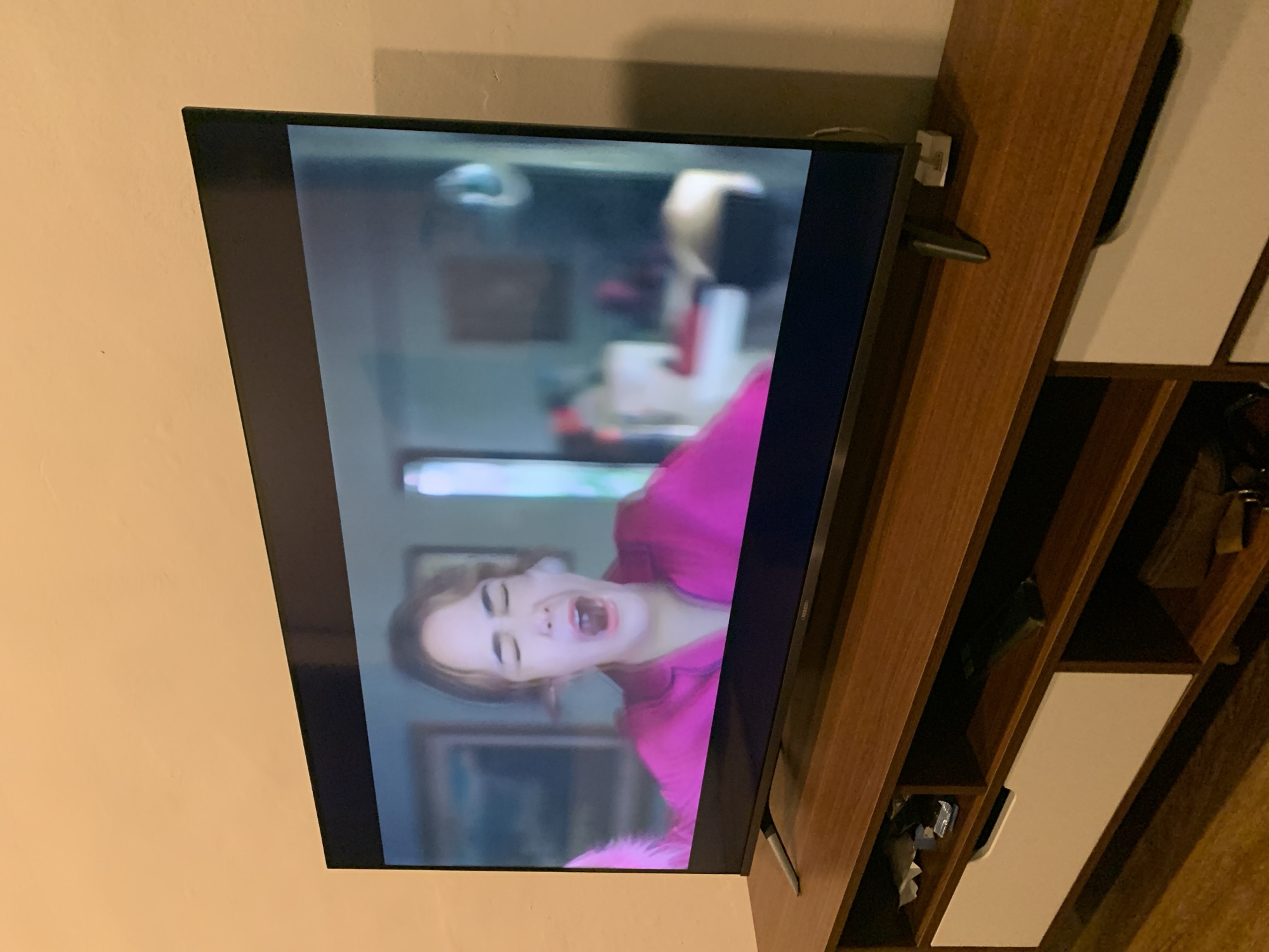 An indoor, eye-level shot shows a person with their mouth open and eyes closed on a television screen, wearing a bright pink top. The television is mounted on a brown wooden entertainment center with white drawers and shelves. The entertainment center is placed against a light orange wall. The overall scene is dimly lit. The image does not provide any specific details about the location in Kualadua, Indonesia.
