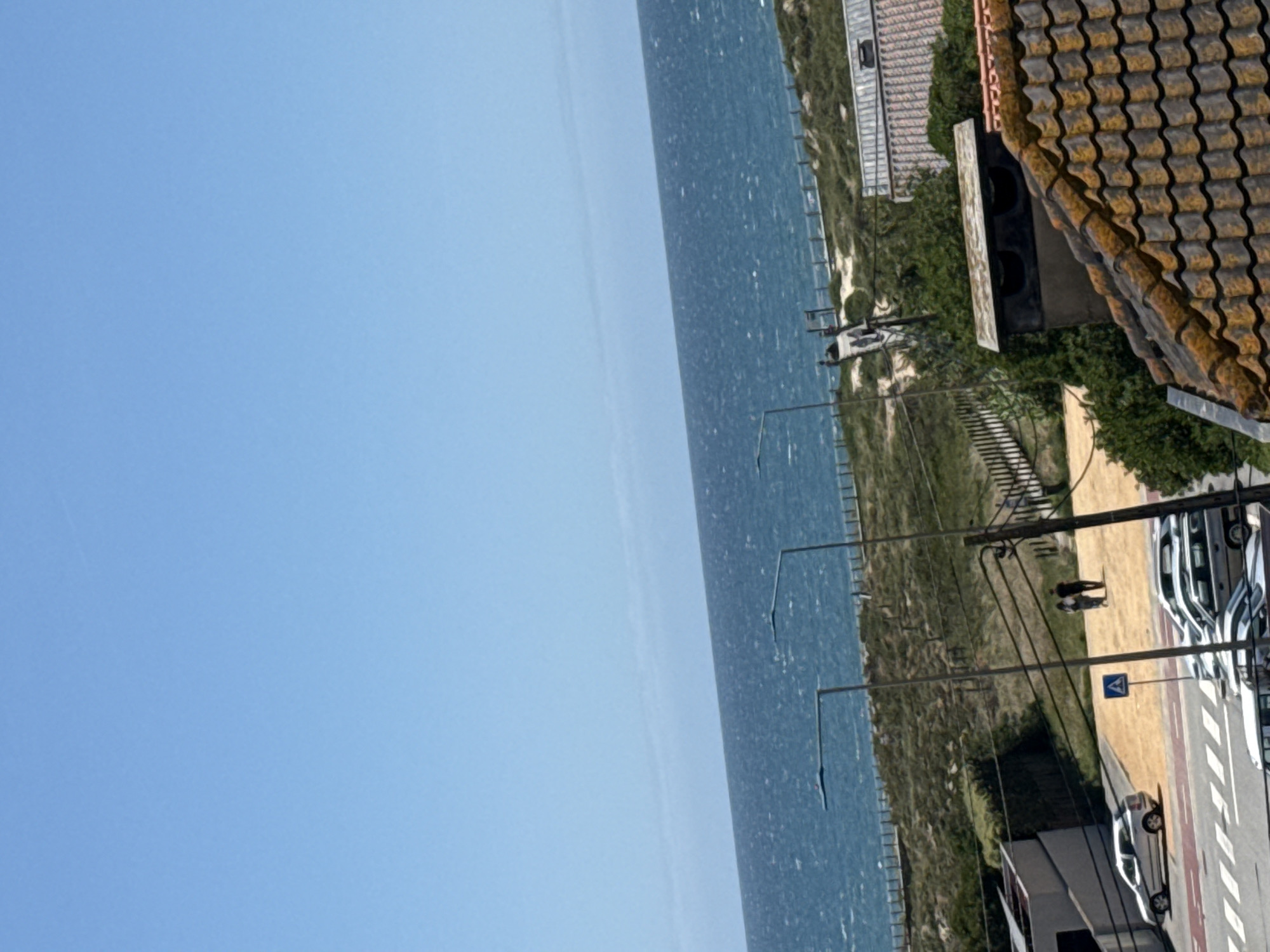 The image presents a view of Vila Nova de Gaia, Portugal, capturing a coastline, residential areas, and street activity. Predominantly, the sky occupies the left portion of the frame, transitioning into a body of water. Along the coastline, there is a sandy strip and low-lying vegetation. A street runs parallel to the coast, where multiple cars are parked in a line, and a person is walking. There is a building with a terracotta-tiled roof visible. Streetlights and road signs are also present.