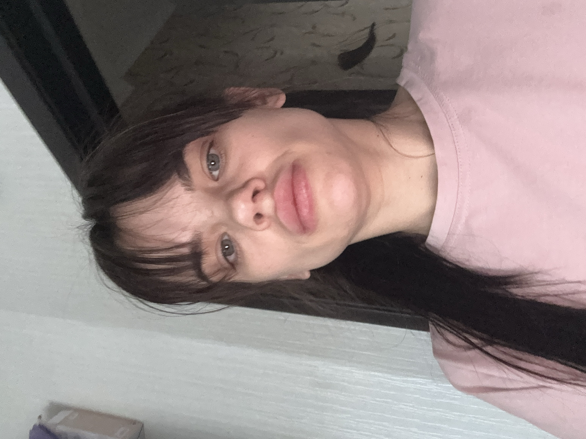A close-up, rotated portrait shows a young woman indoors, likely in Rostov-on-Don, Russia. She has long, dark hair, some of which forms bangs across her forehead, and light-colored eyes. Her lips are full, and she has a slightly pouting or "duck face" expression. She is wearing a simple, light pink t-shirt.
The image appears to be a casual selfie. In the background, a subtle, textured wallpaper with a repeating pattern is visible, suggesting a domestic setting such as a room in a house or apartment. A dark, possibly wooden, structure like a door frame or furniture edge runs along the left and top side of the frame. In the upper background, on the patterned wall, a dark, elongated, curved object, possibly a decorative element or a shadow, is present. A small, light-colored box with a hint of purple is partially visible in the bottom left corner. The lighting is diffused, consistent with indoor conditions, and no specific time of day can be determined. There is no visible text in the image.
