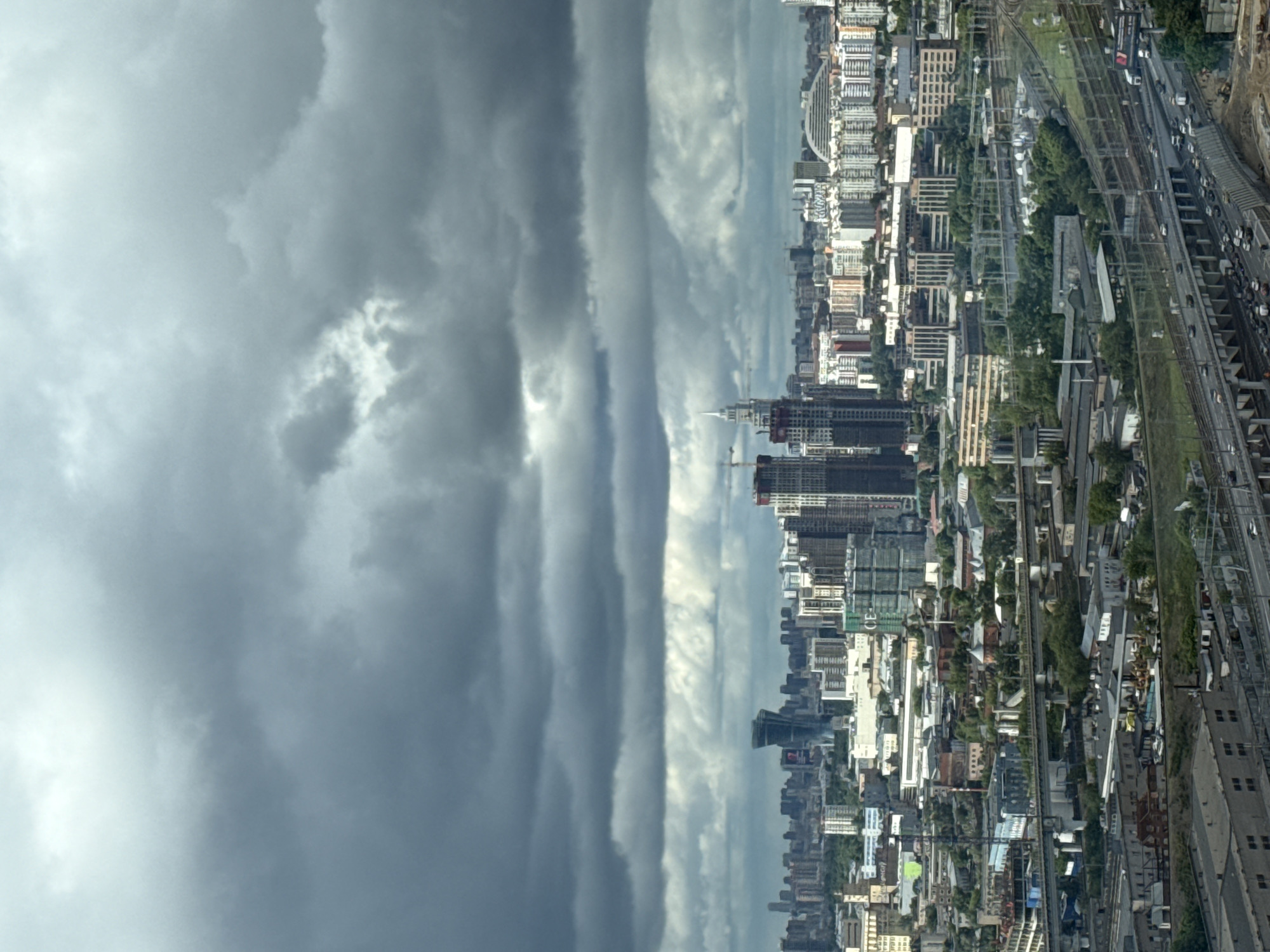 The image shows a cityscape under a stormy sky. The urban area is densely populated with buildings, including skyscrapers and smaller structures, interspersed with green spaces. A railway line runs along one side of the city, with visible trains and traffic. The location appears to be Khimki, Russia, judging by the architectural style and urban layout. The weather is overcast, with dark, heavy clouds dominating the sky, suggesting an impending storm. There are no people clearly visible in the image.