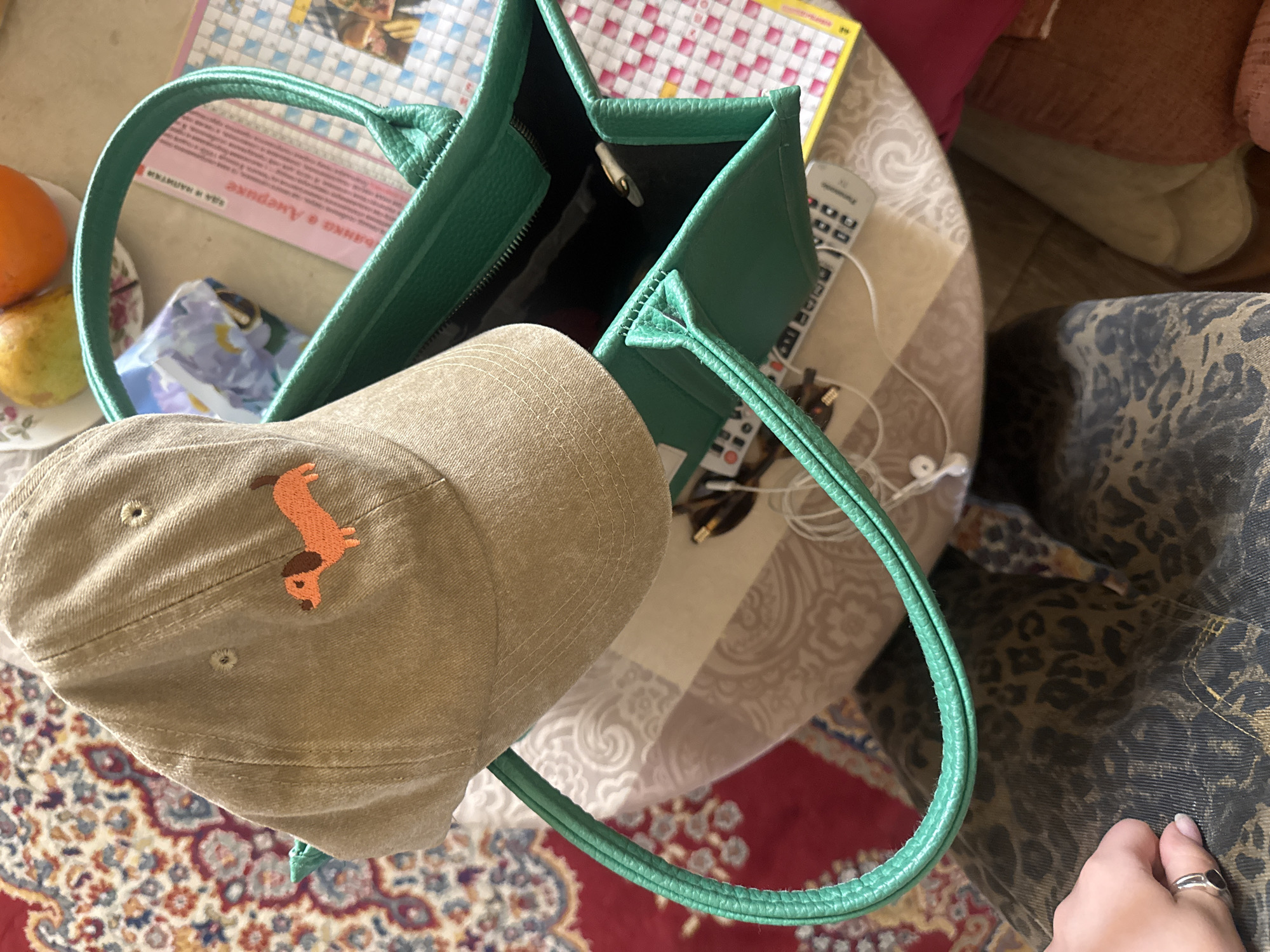 The image presents a high-angle view of a small, round table covered with a patterned tablecloth. A green leather tote bag with black interior is central to the composition, with a khaki-colored baseball cap adorned with an orange Dachshund dog embroidered on the front placed partially inside. An orange and a pear are positioned on a floral-patterned plate adjacent to the bag. Visible within the bag are a set of headphones and a remote control. A crossword puzzle book is also on the table. A small part of a patterned armchair or clothing is visible. A hand wearing a ring is on the lower-right corner. A rug with Persian design is below the table. There is no evidence of location within Yerevan, Armenia.