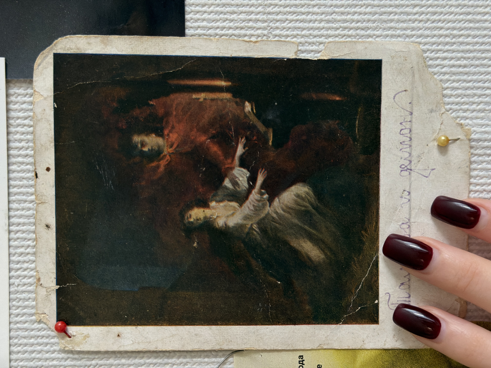 A photograph captures an old, worn print of a dramatic painting, secured to a textured white surface. The central subject of the print is a dark, classical-style painting depicting two figures in motion. One figure, draped in reddish-brown cloth, appears to be falling or is being pulled downwards. The second figure, in lighter, possibly white or pale grey garments, reaches upwards or struggles, their arms extended in distress. The setting of the painting is dark and indeterminate, conveying a sense of struggle or tragedy.
The print itself is a physical object, likely a postcard or a small art reproduction, showing significant signs of age and wear. Its edges are tattered and curled, with visible creases and small tears across its surface. A red thumbtack holds the bottom left corner, and a yellow thumbtack secures the top right corner to the background, which is a coarse, white, woven texture, possibly a fabric wall covering or a bulletin board.
A human hand, with nails polished in a dark red (maroon) color, partially covers the right side of the print and the yellow thumbtack. On the worn white border of the print, handwritten text in purple ink is visible vertically along the right edge, appearing to read "Геннадий" (Gennady) and a numeral "4". Fragments of other Cyrillic text, such as "ода", are visible on another piece of paper partially underneath the main print at the bottom.
The image documents this art print and its display in Zasechnoye, Russia. No specific time of day or weather elements are discernible. The scene focuses on the aged print and its presentation, suggesting it is a personal item.