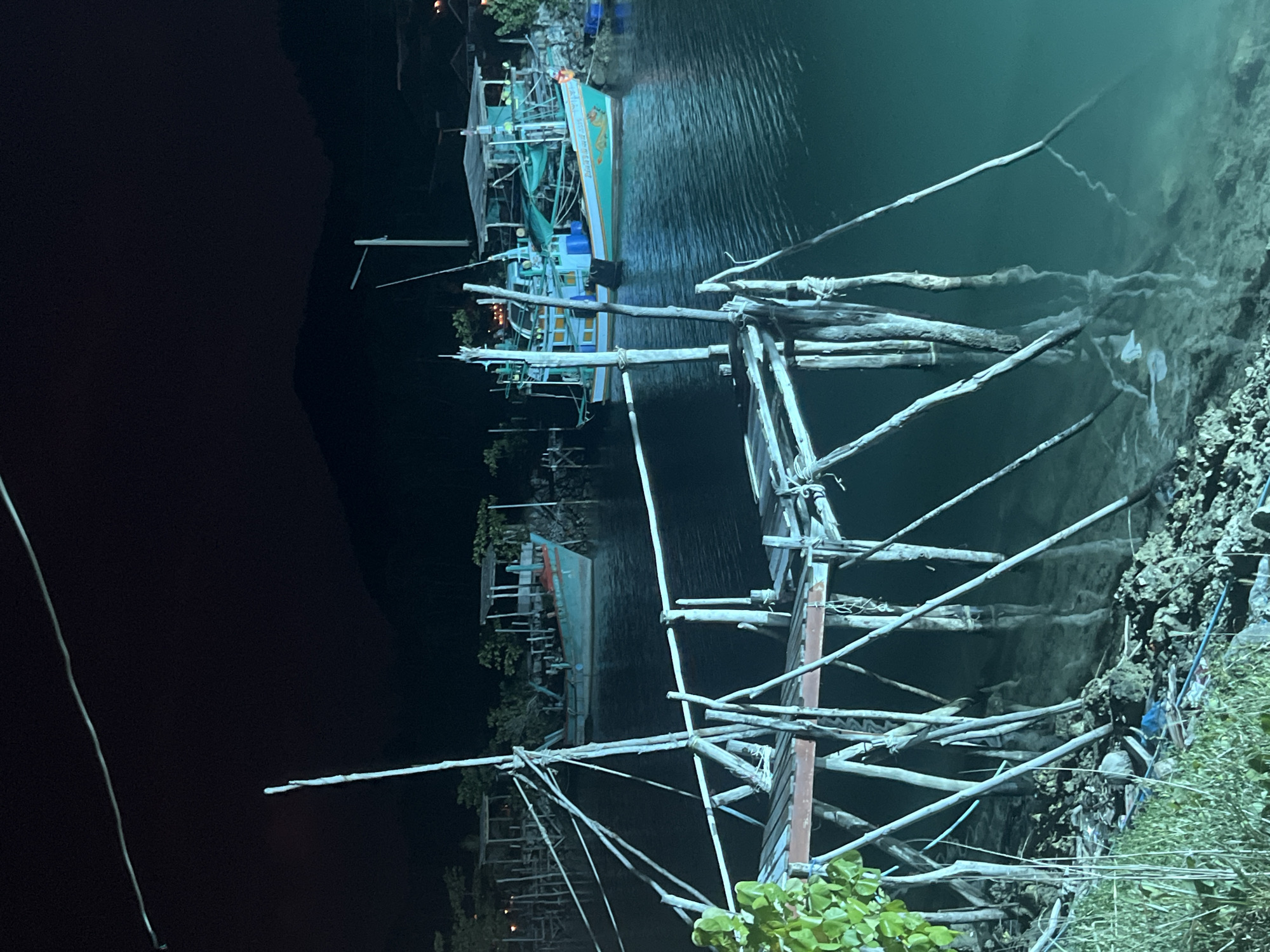 This is a nighttime photograph of a coastal scene in the Ko Pha-ngan District of Thailand. In the foreground, a dilapidated wooden pier juts out into the water, supported by numerous pilings. The pier is constructed with weathered wooden planks and beams, and ropes are visible attached to some of the supports. To the left of the pier, lush green foliage grows along the rocky shoreline.
The water is a deep, dark turquoise, with subtle reflections of the pier and surrounding structures. In the middle ground, two fishing boats are moored. The boat on the right is larger and more brightly lit, with turquoise and white paint and some visible lettering on its hull. The boat on the left is smaller and appears to be of a similar design. Both boats are typical of local fishing vessels in the region.
In the background, a silhouette of dark, tree-covered mountains rises against the night sky. Small, distant lights suggest structures or buildings on the shore. The overall atmosphere is quiet and serene, illuminated by artificial lights from the boats and possibly from the shore, creating a contrast with the dark natural surroundings. The scene depicts a typical working harbor or coastline at night.