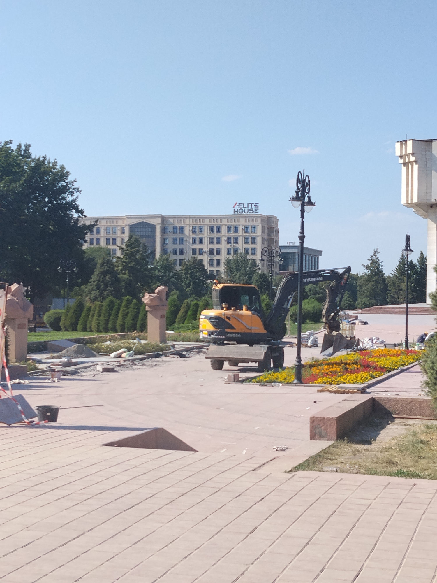 In Bishkek City, Kyrgyzstan, a sunny day reveals a construction site in progress. An excavator is actively working on what appears to be landscaping or renovation of a public area. In the background, a large, modern building with "ELITE HOUSE" signage stands against a clear blue sky. The foreground shows a paved walkway with decorative flowerbeds, some of which have been disturbed, suggesting recent work. Several statues or busts are visible, some appearing to be under protection or in the process of being moved. Construction materials, including sand and bricks, are scattered around the site. Two individuals are present in the background, seemingly engaged in the construction activities. The overall scene depicts a public space undergoing significant refurbishment.
