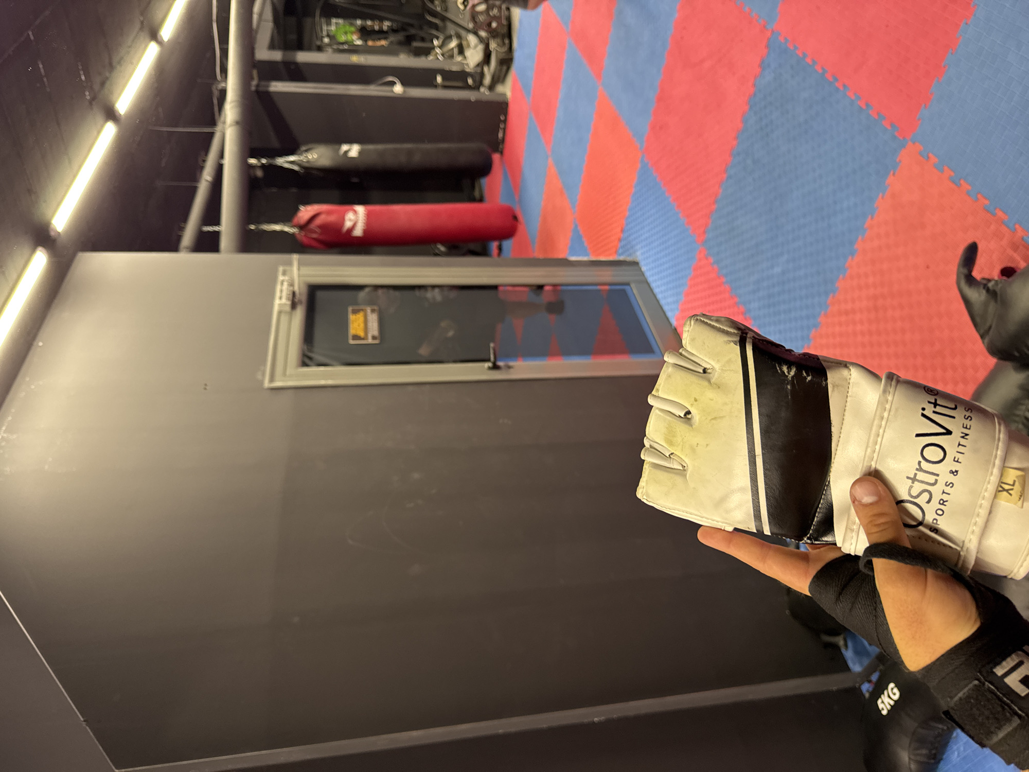 A first-person perspective captures a person's right hand and forearm, equipped for training in what appears to be a gym or martial arts studio. The hand wears a white and black fingerless MMA glove, showing signs of use and dirt, with "OstroVit SPORTS & FITNESS" and "XL" printed on it. Beneath the glove, a black wrist wrap with a hook-and-loop closure is visible around the wrist.
The setting features a prominent dark grey or black wall on the left and foreground, which includes a rectangular, framed opening or window reflecting dark interior elements. Above this wall, linear fluorescent lights illuminate the ceiling. In the background, two long, cylindrical punching bags, one black and one red, hang from a rack or ceiling fixture. The floor is covered with interlocking foam mats arranged in a checkered pattern of blue and red squares, typical of combat sports training areas. Part of a black object with "5KG" visible on it lies on the floor near the bottom right. The overall impression is an indoor training environment for boxing or martial arts.