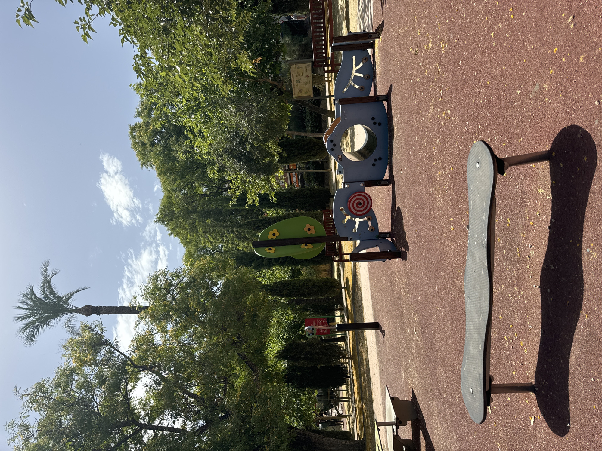 The image depicts a playground scene on a sunny day. In the foreground, a playground structure with blue and green elements is visible. One blue section features a large circular opening and a red and white spiral design. Attached to it is a green, stylized tree shape with yellow flower-like accents. To the right, a dark gray, curved object, possibly part of another playground apparatus, casts a long shadow on the reddish-brown rubberized ground.
In the background, lush green trees create a canopy, with a tall palm tree visible on the left side of the frame. The sky is a clear blue with a few scattered white clouds. The sun is high, indicating it is likely midday. The overall impression is one of a bright and pleasant outdoor environment, typical of a park or recreational area. No people are visible in the image. There is some faint text on a red sign near the center of the image, but it is too blurry to be deciphered. The presence of a palm tree and the clear blue sky suggest a warm climate, consistent with the location context of Murcia, Spain.