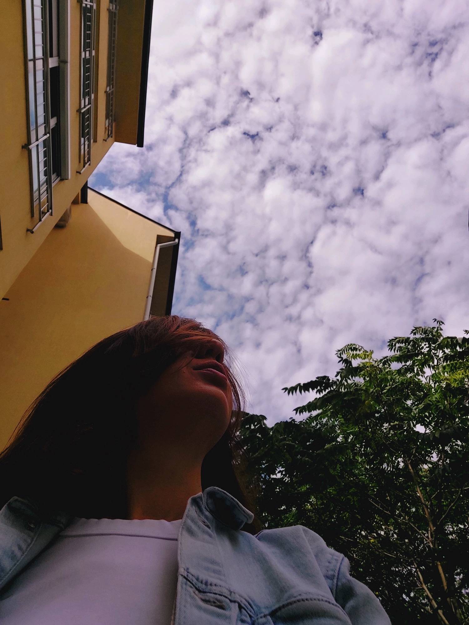 An eye-level medium shot captures a young person, possibly female, from the chest up, looking upwards. They are wearing a light blue denim jacket over a white t-shirt. Their head is tilted back, with dark hair falling across their face and obscuring their features. The background is a mix of urban elements and nature, with a pale yellow building on the left and lush green trees on the right. Above, a cloudy sky dominates the frame, with scattered white clouds against a light blue backdrop. The building, with its repeating windows and balconies, suggests an apartment block or office building. The perspective is from a low angle, looking up towards the sky and the upper portions of the structures and foliage, implying the photo was taken from ground level. The lighting is soft and diffused, likely due to the overcast sky, creating a somewhat muted color palette. The scene appears to be outdoors in a city environment, possibly in Sofia, Bulgaria, given the architectural style of the building and the presence of trees.