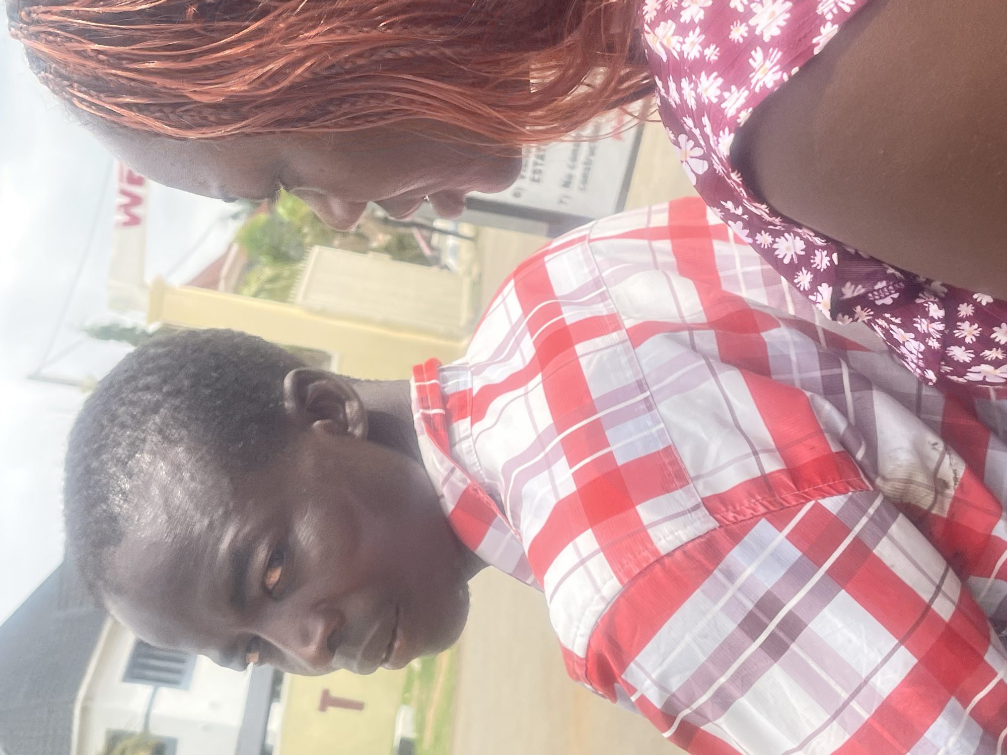 Two individuals are closely positioned outdoors. One person, wearing a red and white plaid shirt, is facing forward with a slight tilt to the left. The second person, seen partially from above, has braided hair with reddish tones and is wearing a floral print top. The background includes buildings and greenery. A sign with the letters "WE" is partially visible. Based on the available context, this scene appears to be in a residential or commercial area. Further information is needed to identify the exact location in Abuja, Nigeria.