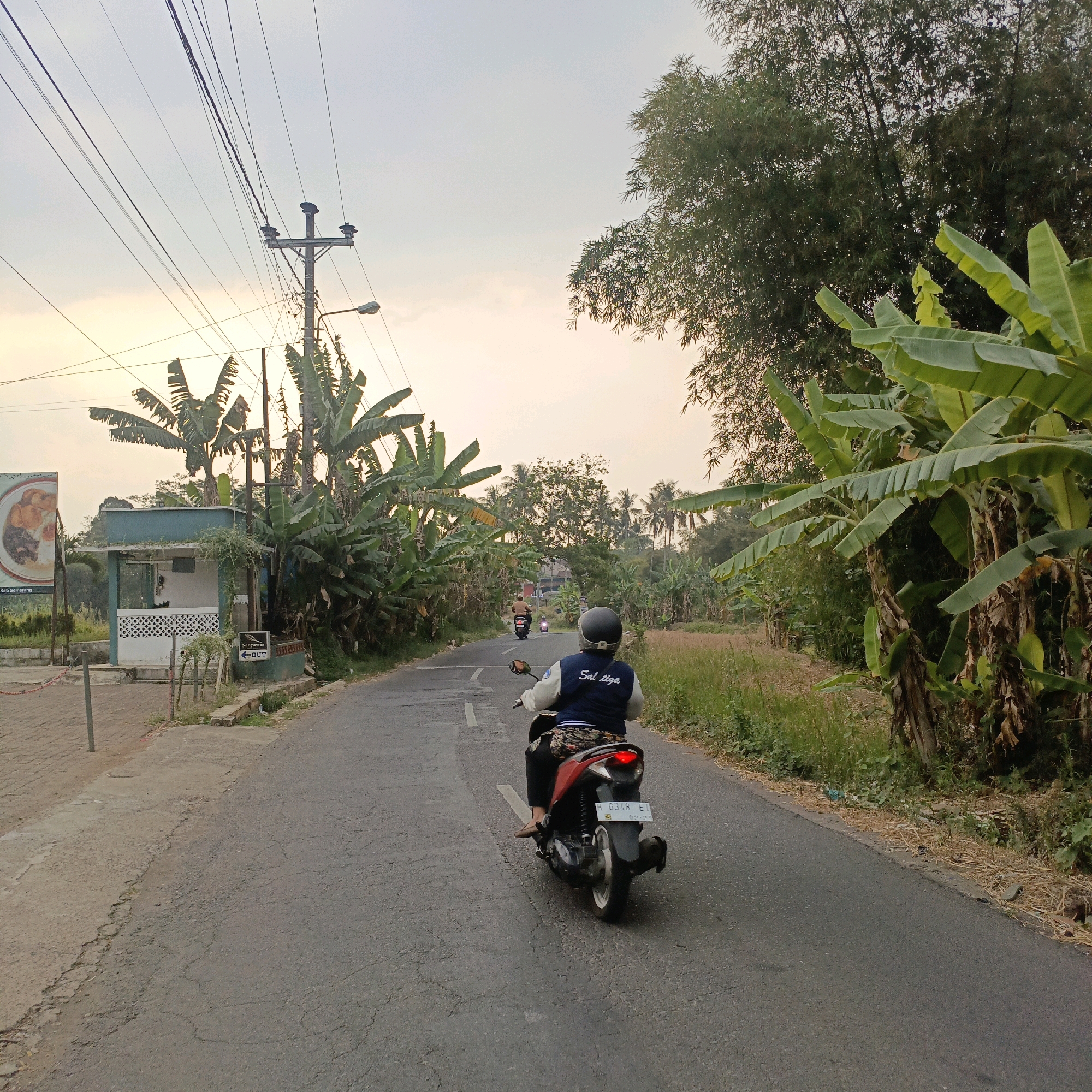 The image depicts a street scene in Jombor, Indonesia, with a clear view of banana trees and bamboo vegetation lining the right side of the road. The sky is overcast with a yellowish hue, suggesting it might be dusk or dawn. In the foreground, a person is riding a red motorcycle, wearing a dark blue vest with white lettering that appears to read "Sal tiga". The motorcycle has a license plate that reads "H 6348 EI". The road is paved and curves slightly to the left. On the left side of the road, there is a small building with a teal-colored roof and white walls, possibly a kiosk or small shop. A sign with a picture of food is visible on the left. A utility pole with many wires runs along the left side of the road, accompanied by a street lamp. Further down the road, two more motorcycles are visible, one ahead of the rider in the foreground and another further in the distance. The overall impression is a rural or semi-rural setting within Jombor.