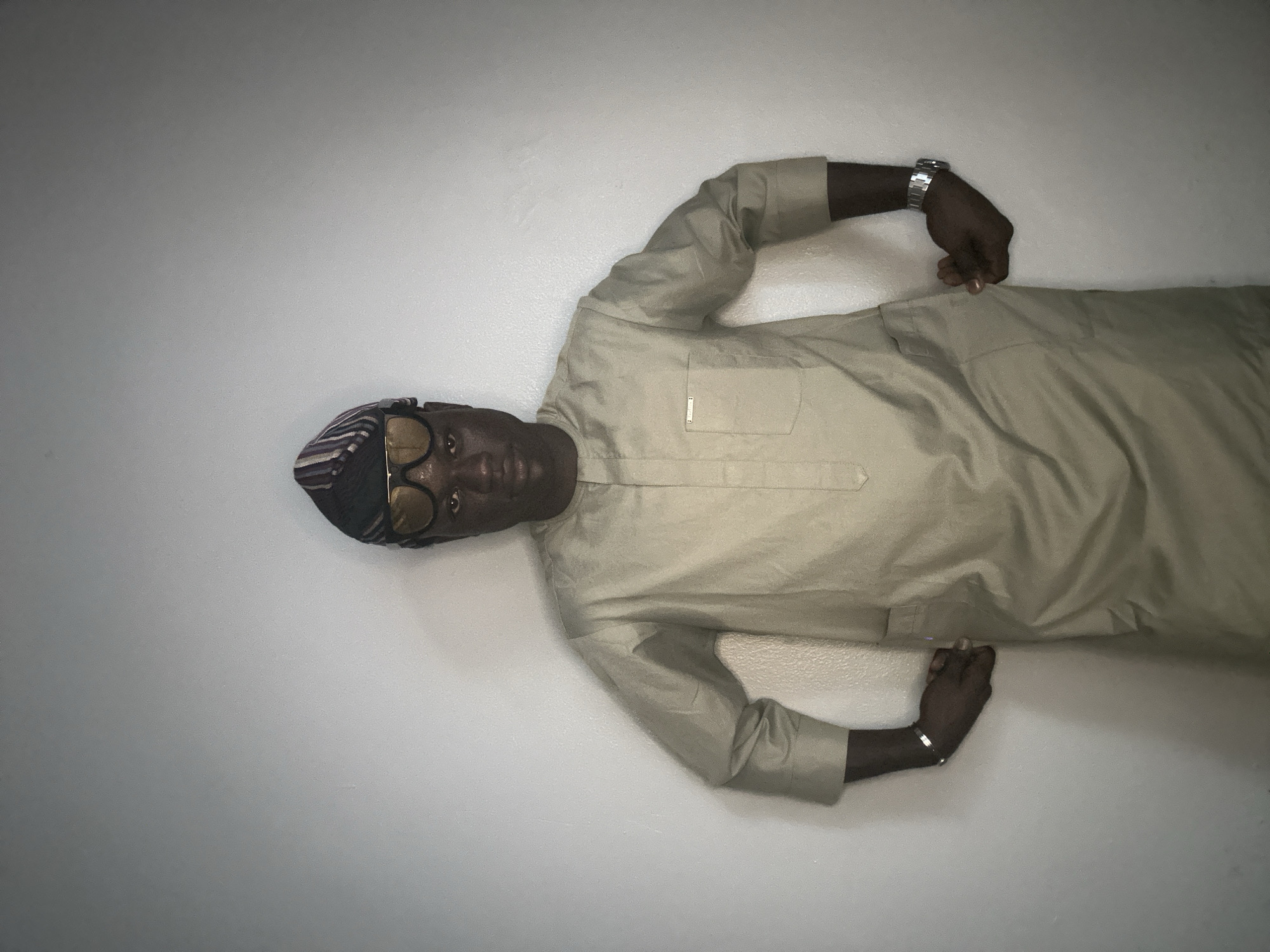 A man stands against a plain white wall, wearing a light tan, long-sleeved garment with a mandarin collar and a breast pocket. He also wears a patterned, brimless cap and large, tinted sunglasses. His hands are placed inside the pockets of his garment. A silver watch is visible on his left wrist. This image was captured in the city of Badagry, Nigeria.