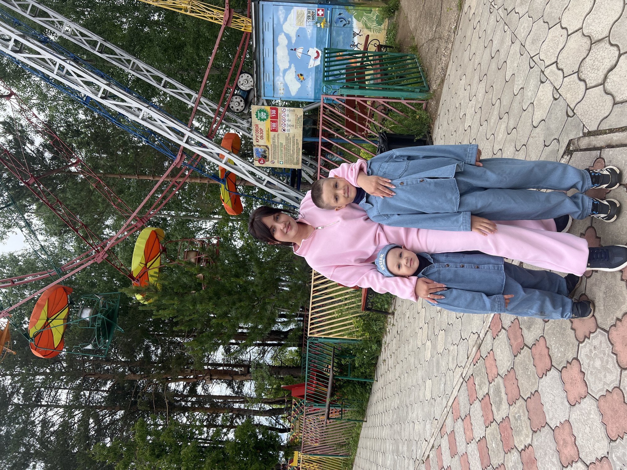 An outdoor scene in an amusement park in Chaikovsky, Perm Krai, Russia, features three individuals posing for a photograph. The individuals consist of an adult female and two male children. The woman, appearing to be in her late 20s or early 30s with dark hair, is wearing a light pink tracksuit comprising a high-collar zip-up top and matching pants. She has her right hand resting on the left shoulder of the older boy and exhibits a slight smile.
To the woman's right stands an older boy, approximately 6-8 years old, with short brown hair. He is dressed in a light blue denim-style long-sleeved shirt over a dark turtleneck and matching light blue denim-style pants, paired with black and white athletic shoes. He also presents a slight smile. To the woman's left is a younger boy, approximately 2-4 years old, with short brown hair, wearing a similar light blue denim-style long-sleeved shirt and pants, black athletic shoes, and a blue baseball cap embroidered with "BOSTON" in white text. He looks towards the camera with a neutral expression.
The background is dominated by a large, multicolored Ferris wheel featuring red, white, blue, and yellow structural elements. Its cabins are green with orange and yellow sunshade coverings. Dense tall pine trees and other deciduous foliage are visible behind and around the Ferris wheel. To the right of the scene, a small, light blue wooden building functions as an operator's booth or ticket kiosk, adorned with a mural depicting a beach scene with a sailboat, fish, and palm trees. A white sign affixed to the Ferris wheel structure or an adjacent post provides ride information in Cyrillic, including "КРУГОВОЙ ОБЗОР" (Panoramic View), pricing (180 and 130 units), "ОДИН КРУГ" (One Ride), and safety regulations. A metal fence, painted in sections of green, red, and yellow, runs horizontally across the midground, behind which are benches and overgrown vegetation. Several wooden birdhouses are attached to the pine trees.
The ground in the immediate foreground is paved with interlocking concrete pavers. The left side features light pink/reddish cloud-shaped pavers embedded in a lighter gray pattern, while the right side displays a wavy, interlocking light gray pattern. The sky is overcast, providing diffuse lighting for the scene. The presence of Cyrillic text on the sign and the regional code "59" (for Perm Krai) visible on the building confirm the location as Chaikovsky, Russia.