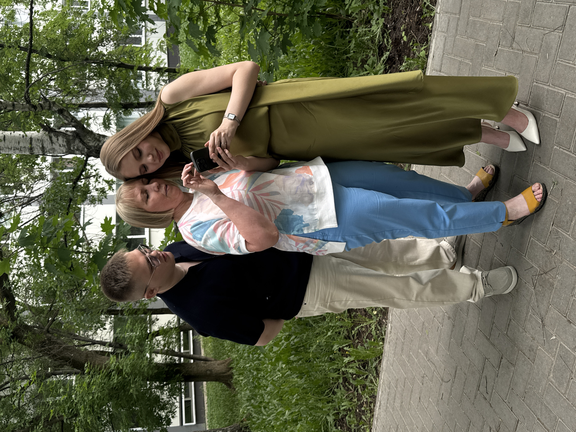 Three people are standing on a paved walkway near a grassy area and trees. A woman in a long olive dress and white heels is holding a cell phone and leaning her head close to another woman wearing a floral shirt and blue pants, who is also looking at a cell phone. A man in a black shirt, khaki pants, glasses, and grey shoes is standing below them. The background includes trees, a building, and foliage, suggesting an urban park setting. Based on the architecture and vegetation, it is likely a location in the city of Perm, Russia.
