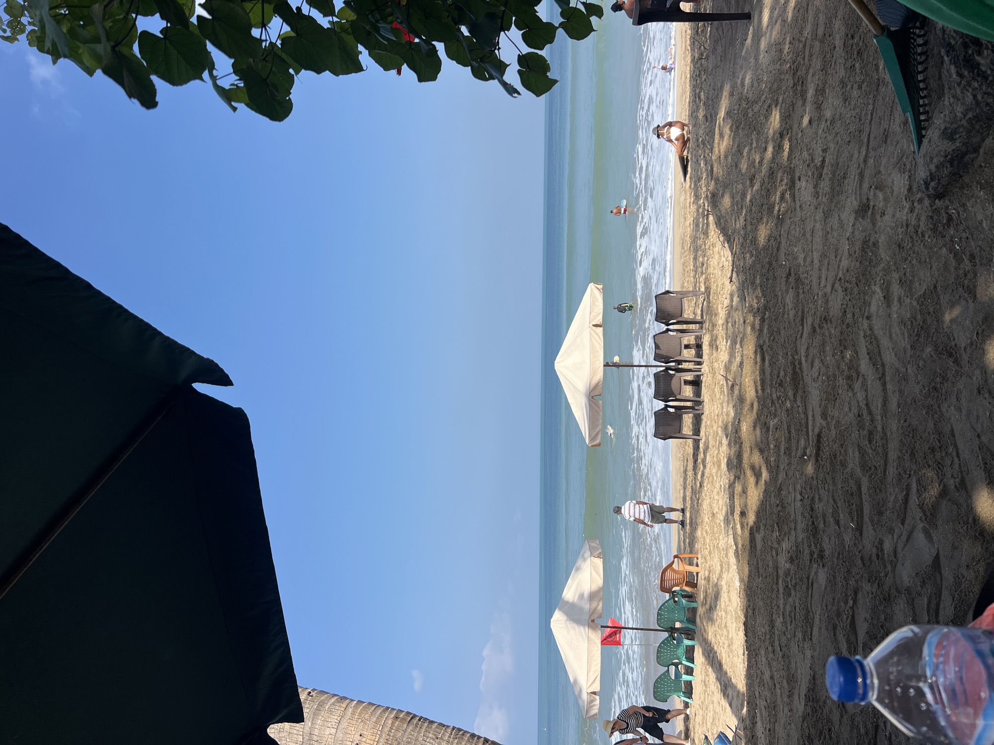 In Kuta, Indonesia, a beach scene unfolds under a clear blue sky. Two large, light-colored beach umbrellas provide shade on the sand. In the foreground, a plastic water bottle with a blue cap is visible. Several empty plastic chairs, in various colors including green and orange, are arranged in front of the umbrellas. A red flag is planted in the sand near one of the umbrellas. Several individuals are present. Two people stand near the water's edge, facing the ocean. Another person is seated on the sand, facing the water, wearing a white bikini and a hat. In the ocean, several people are swimming or wading. The sea has gentle waves. The image is framed by the dark canopy of another umbrella in the upper left and green foliage on the right. The sand is textured with footprints and debris.
