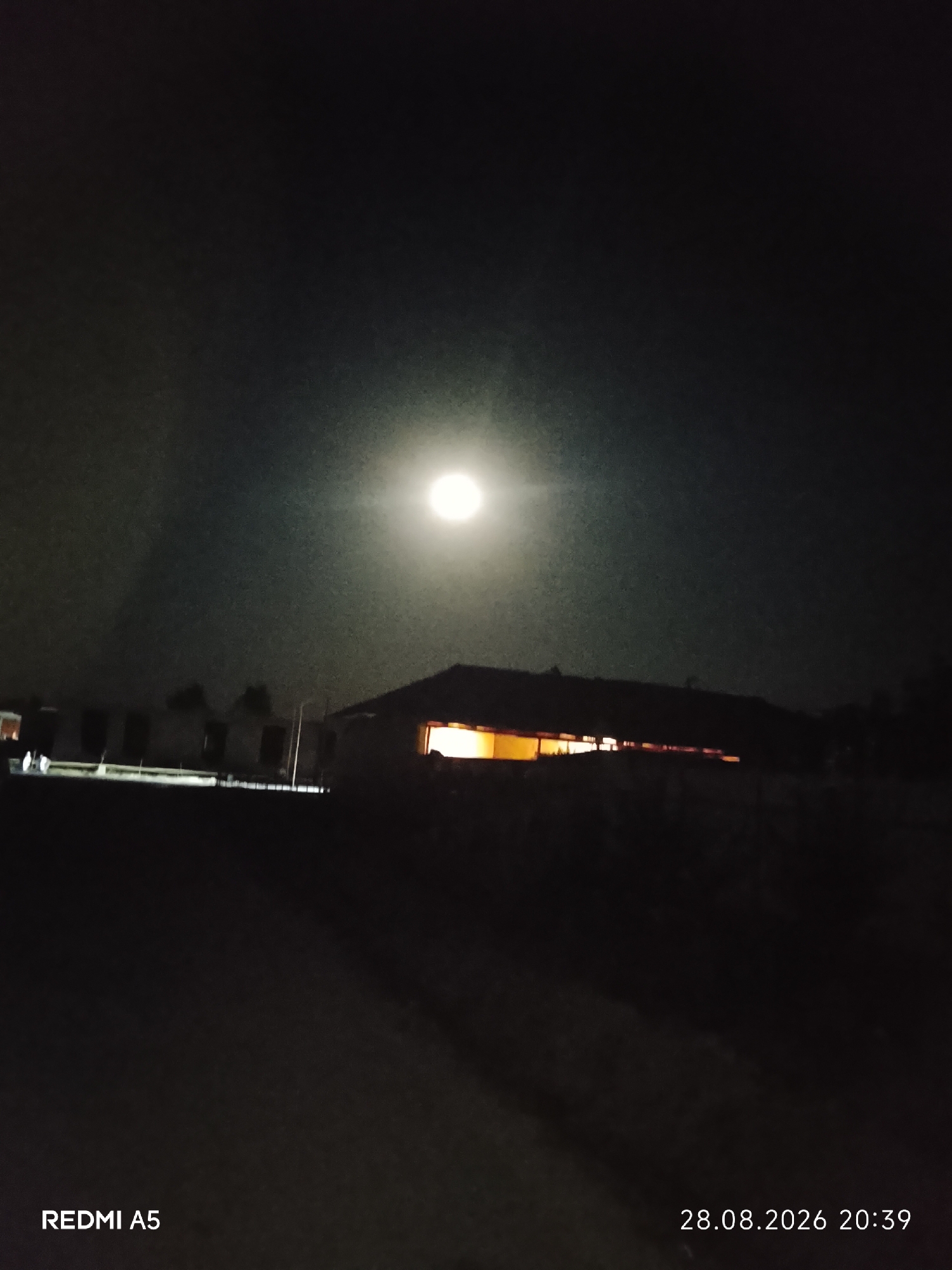 A night-time outdoor scene captures a brightly lit full moon in a dark, hazy sky. In the mid-ground, a low-lying building with illuminated windows is silhouetted against the sky. In the foreground, the ground appears to be a dark, unpaved surface. The bottom of the image displays text "REDMI A5" on the left and "28.08.2026 20:39" on the right. The location is identified as Pakhtakor, Uzbekistan. There are no people or active interactions visible. The environment is still and quiet, with the dominant feature being the moonlit landscape.