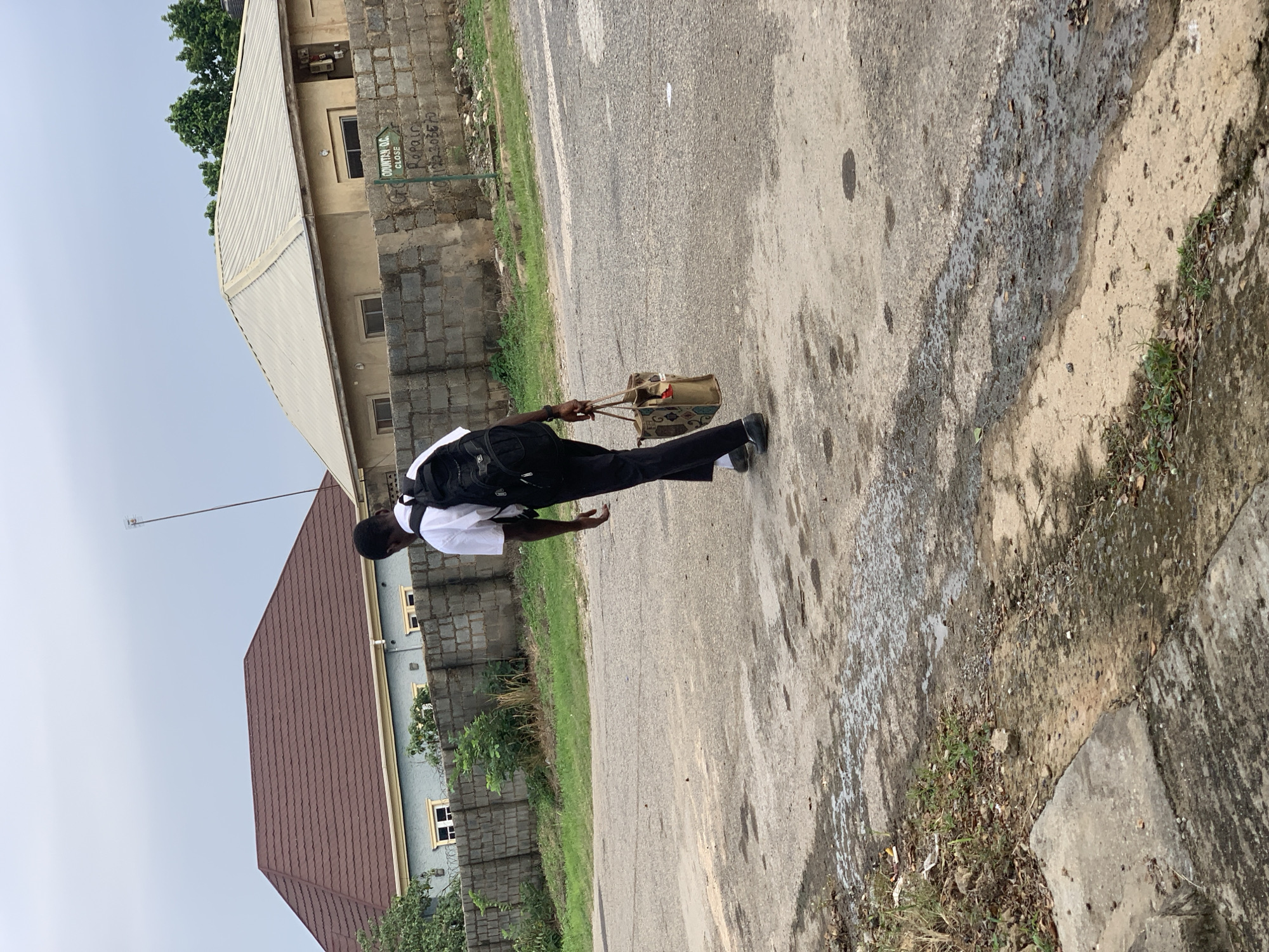 A young Black male wearing a white shirt, black trousers, black shoes, and a black backpack walks away from the camera down a paved road in an outdoor urban environment. He is carrying a tan bag with a red pocket in his right hand. The road is wet and appears to be partially paved with some debris on the side. In the background, there are two single-story buildings with brick walls and tiled roofs. One building is light brown, and the other is painted light blue with a brown roof. A low brick wall separates the buildings from the road. A green sign with white lettering is attached to the brick wall. Trees are visible behind the buildings. The sky is overcast.