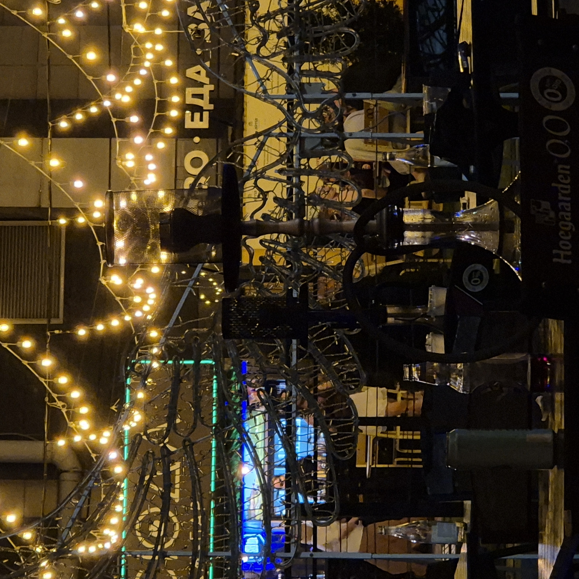 The image depicts an outdoor seating area at night, likely a cafe or bar, in Kemerovo, Russia. The scene is illuminated by strings of warm yellow fairy lights draped across the foreground and over a complex metal structure. A prominent hookah pipe stands in the mid-ground, with its base on a black stand that has some visible branding, including "Hoegaarden 0.0%".
In the background, a sign with Russian text is visible, featuring the letters "О ЕДА". Blue and green neon lights are also present in the background, possibly indicating a display or advertisement. Several tables and chairs are set up, with some patrons visible through a partially obscured barrier. The overall atmosphere is dimly lit, with the fairy lights providing the primary illumination. Several glass bottles, including a clear bottle and a dark-colored bottle, are placed on tables. The metal structure in the foreground appears to be decorative or part of the venue's facade, with intricate curved elements.