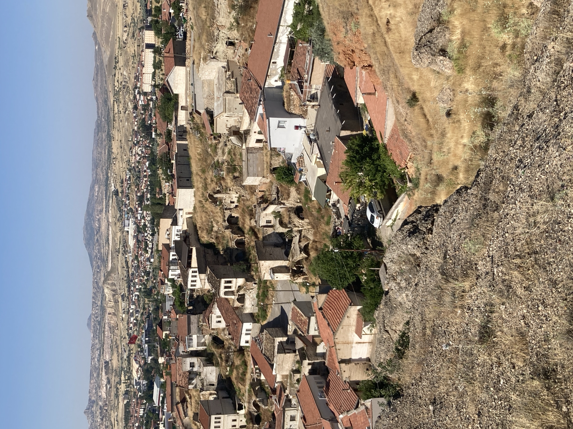 This is a high-angle, medium shot of Avanos İlçe Merkezi, Turkey. The image captures a dense cluster of buildings with red-tiled roofs, nestled into a hilly, arid landscape. Many of the structures appear to be integrated into the natural rock formations, with some exhibiting cave-like entrances. The architecture suggests a blend of traditional and more modern construction, with varying materials like stone and plaster evident. Dry, yellowish-brown grass and scattered rocks cover the terrain, indicating a warm, dry climate. In the distance, a wider view of the city reveals more buildings and the undulating, rocky topography of the region, with a clear blue sky overhead. A Turkish flag is visible on a flagpole in the middle ground, adding a national marker to the scene. A white car is parked on a road winding through the settlement. There are no people prominently visible in the frame.