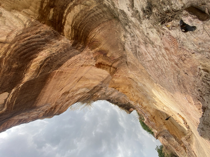 This image, captured in Amangwu, Nigeria, presents an upward perspective of a deep, excavated or naturally eroded geological formation. The scene is dominated by towering, stratified earthen walls composed of reddish-brown, orange, and light beige sediments. Prominent vertical striations and undulating horizontal patterns are visible across the wall surfaces, indicative of erosion or excavation. An overcast, cloudy sky is visible at the top. At the lower-left, a single dark-colored sandal rests on a rock. High on the left wall, a small, dark object resembling a bird is clinging to the surface. Sparse dry vegetation is present along the upper ledges and scattered near the base of the walls. No human figures are present.