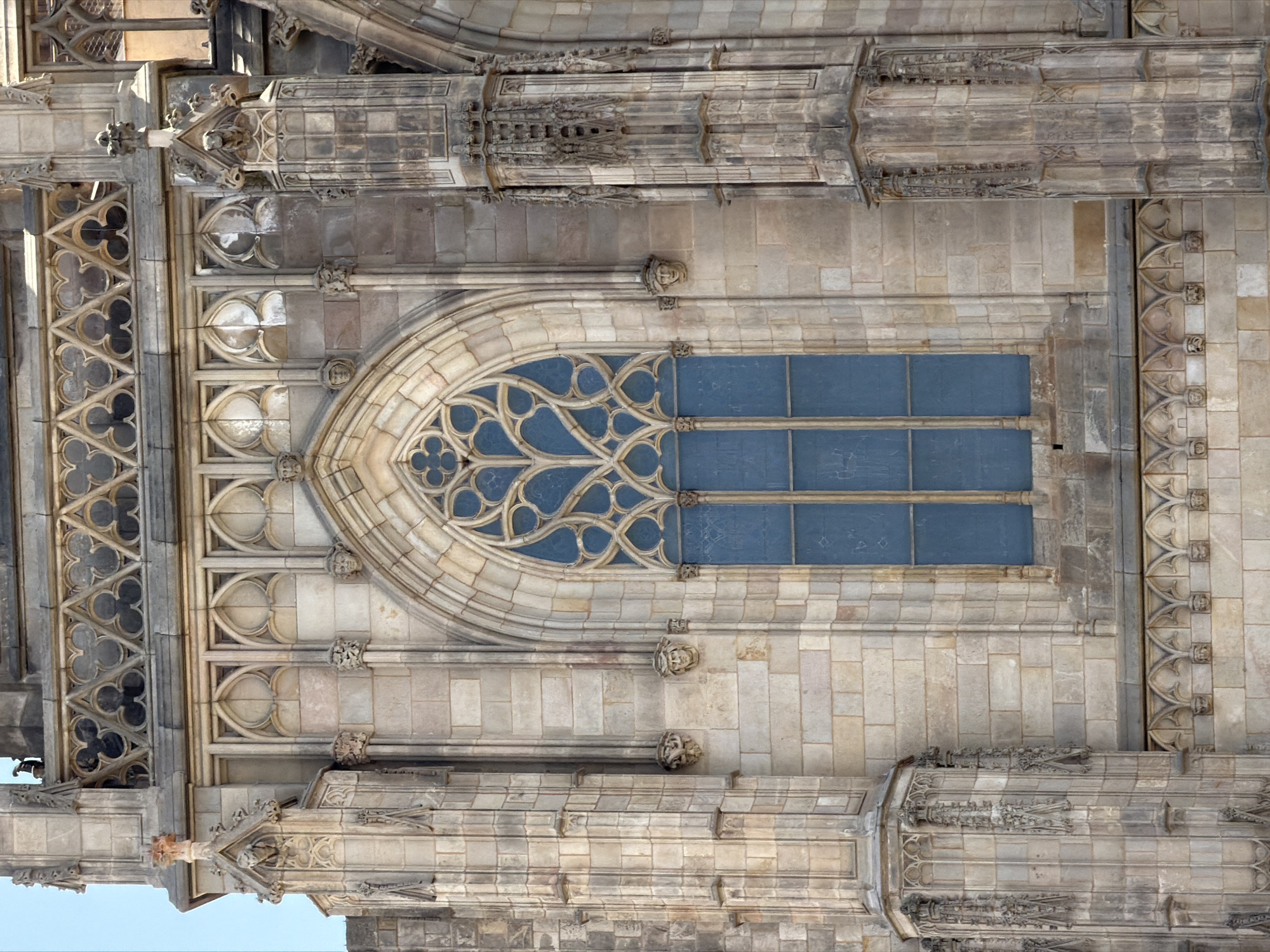 The image shows a detailed view of a Gothic-style window on the exterior of a stone building, likely a cathedral, in Barcelona, Spain. The window is tall and arched, with intricate tracery forming a floral pattern. The glass is a deep blue. The stonework of the building is weathered, with a mix of square and rectangular blocks. Elaborate carvings, including gargoyles and decorative moldings, adorn the facade. The architectural style suggests a historical religious building. The sky is a clear, pale blue, indicating daytime.