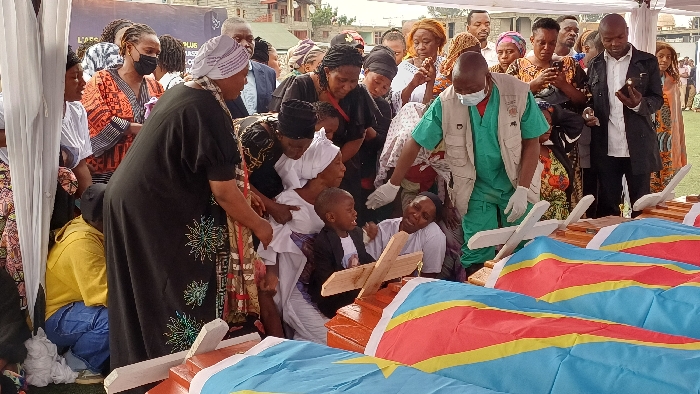 The image depicts a somber outdoor gathering in Goma, Democratic Republic of the Congo, centered around a row of at least six light brown wooden coffins. Each coffin is draped with the national flag of the Democratic Republic of the Congo, featuring a light blue field, a yellow diagonal stripe bordered by red, and a yellow star. Small, simple wooden cross-like markers rest on top of each coffin.
A dense crowd of individuals, predominantly adults of various ages, is gathered around and behind the coffins. Many individuals display expressions of grief or solemnity. Several women wear head coverings, including white and patterned fabrics. One man, positioned near the center-right, wears a light green scrub top, a tan vest, white medical gloves, and a face mask, suggesting a professional or aid worker role. He appears to be observing or attending to the scene. A young boy in a dark jacket is visible among grieving adults near the center-left, with a distressed expression. Several individuals, including a man in a black jacket and white shirt on the far right, are holding smartphones, some appearing to record or photograph the scene.
The immediate environment consists of a grassy area, partially shaded by a white fabric canopy visible on the left. In the background, an urban setting with multiple single and multi-story buildings is visible under an overcast sky. A dark banner with partial white text, "L'ASS" and "PLUS," is also visible in the background.
