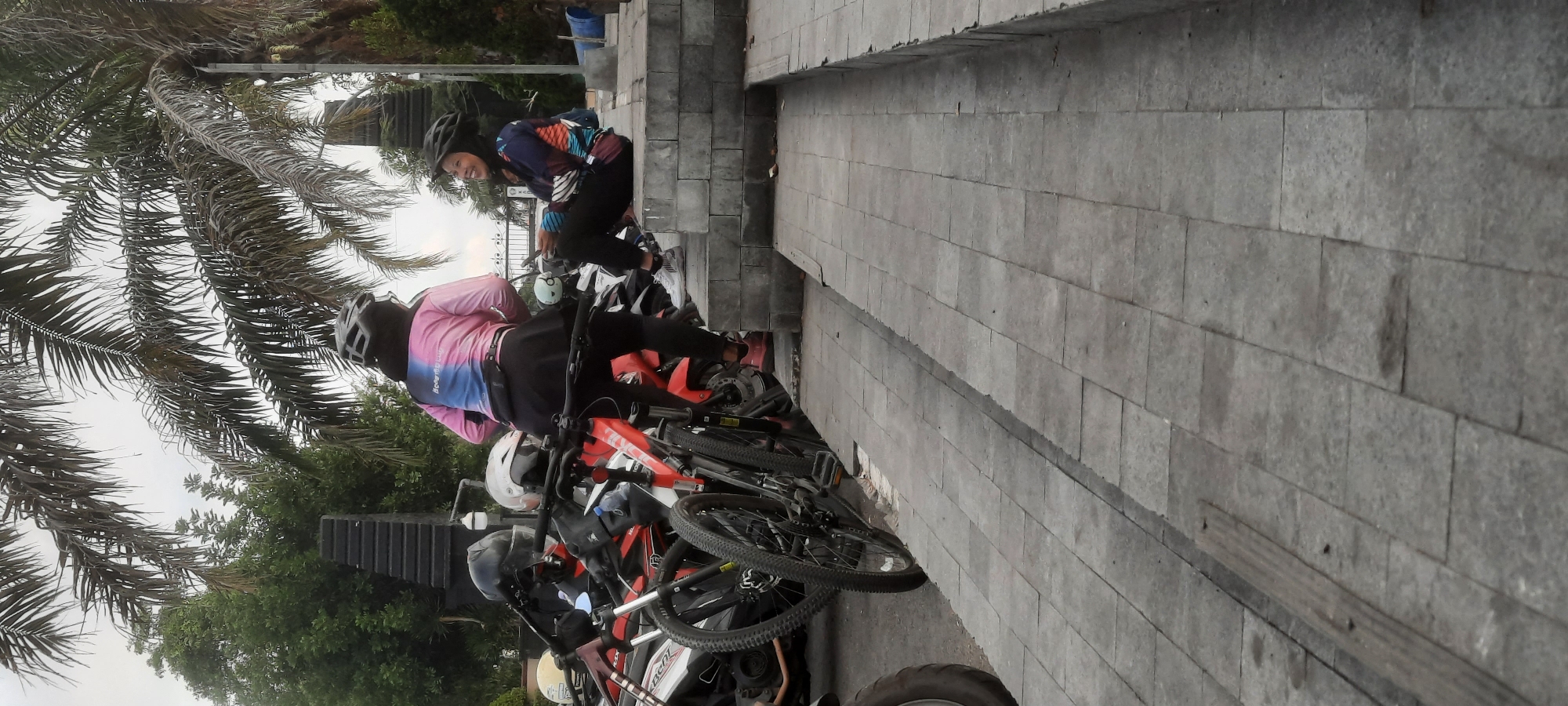 In Kanigoro, Indonesia, two women in cycling attire are present near parked motorcycles. One woman, wearing a pink and blue long-sleeved cycling jersey and black leggings, stands next to a red bicycle, facing another woman. The second woman, dressed in a similarly patterned long-sleeved cycling jersey and black pants, is seated on a concrete step structure. She is smiling and looking at her phone. Both women are wearing cycling helmets. Several motorcycles are parked behind them, with their tires visible. The ground is paved with grey square tiles, and a stone step structure runs along the right side of the image. Palm trees are visible in the background under an overcast sky.