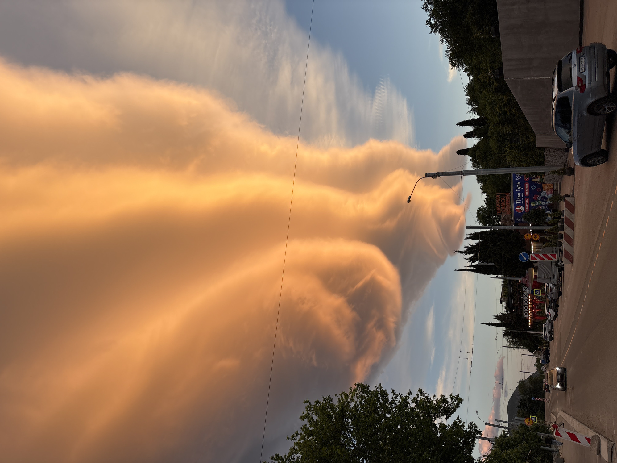 The image shows a street scene in Kiparisnoe, Ukraine. The photo is taken from a low angle. The foreground contains a sidewalk and a road with parked cars and traffic signs. A single car is driving on the road. Trees line the side of the road. In the background, distinctive golden-colored cloud formations are visible in the sky. A utility pole is visible. A building with signage in Ukrainian is present. A parked grey car is in the top right corner.