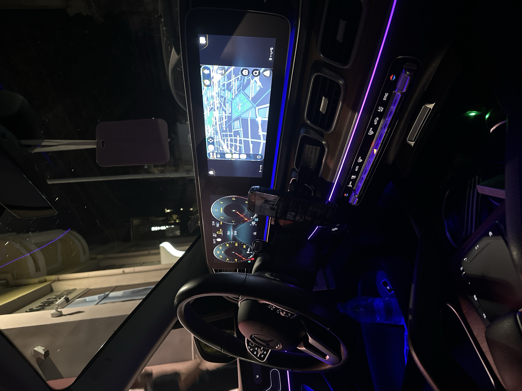 The image captures the interior of a modern car, likely a Mercedes-Benz, at night. The ambient lighting within the car is predominantly a vibrant purple, accentuating the dashboard, steering wheel controls, and air vents. A large, sleek infotainment screen displays a navigation map with a blue and green color scheme, showing a grid-like city layout. To the left of the screen, a digital instrument cluster shows speed, RPM, and other vehicle data. The steering wheel, also illuminated by the purple light, is partially visible on the left side of the frame. A mobile phone is mounted on the steering column. Outside the car, through the windshield, the faint lights of a city street are visible, suggesting an urban environment. The scene appears to be in Ruse, Bulgaria, based on the external context provided. A purple air freshener or charm hangs from the rearview mirror.