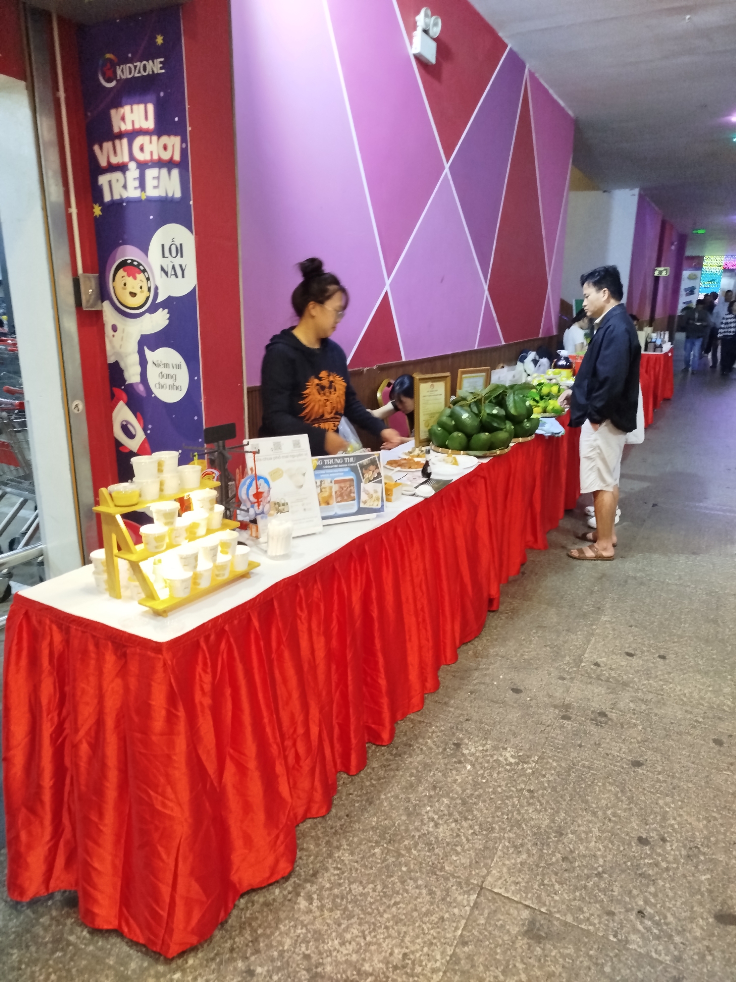 In Da Lat, Vietnam, an indoor scene depicts a brightly lit hallway, possibly within a shopping mall or event venue, characterized by a vibrant wall adorned with a geometric pattern of pink and red triangles. In the foreground, a long table draped in a red tablecloth displays an assortment of items. To the left, a prominent blue banner with a cartoon astronaut and text in Vietnamese announces "KIDZONE KHU VUI CHOI TRE EM," translating to "KIDZONE CHILDREN'S PLAY AREA." Next to it, on the table, is a yellow tiered stand holding numerous white cups. Further down the table, a selection of avocados and other fruits are displayed in a woven basket. A person in a dark jacket and white shorts stands by the table, facing away from the camera. Behind the table, a woman wearing glasses and a black top with an orange lion graphic is serving customers. Additional tables with red coverings are visible further down the hallway, with more people milling about. The floor is tiled with a textured grey material.