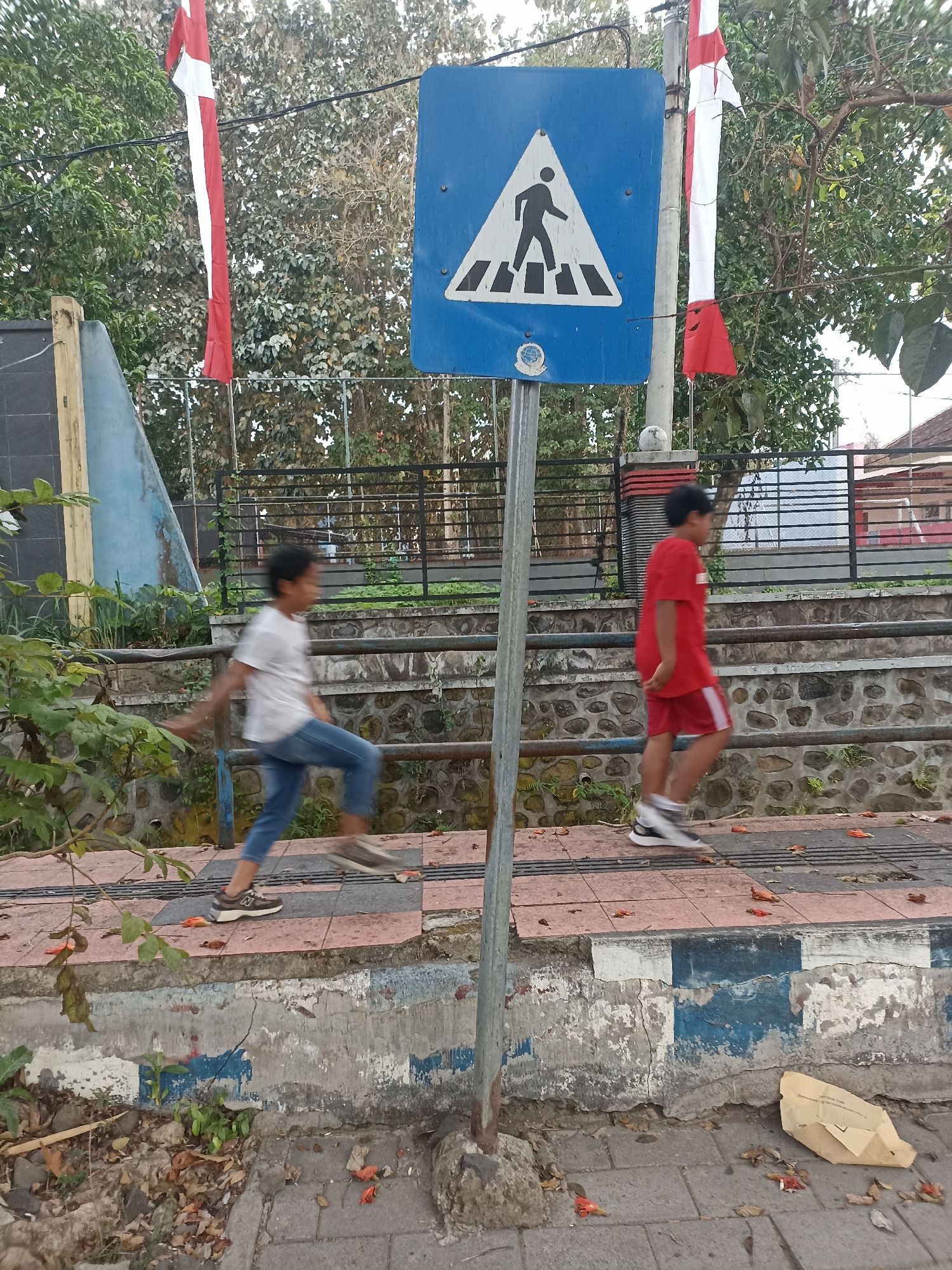 Two young boys are walking on a sidewalk in Mojopanggung, Indonesia. The boy on the left is wearing a white t-shirt and blue jeans, while the boy on the right is wearing a red t-shirt and red shorts. Both boys are in motion, suggesting they are running or walking briskly. A pedestrian crossing sign is visible on a metal pole in the foreground. Red and white Indonesian flags are hanging from wires in the background. The sidewalk is made of patterned tiles, and there is a concrete barrier with blue and white markings. The area appears to be outdoors, with trees and foliage surrounding the scene.