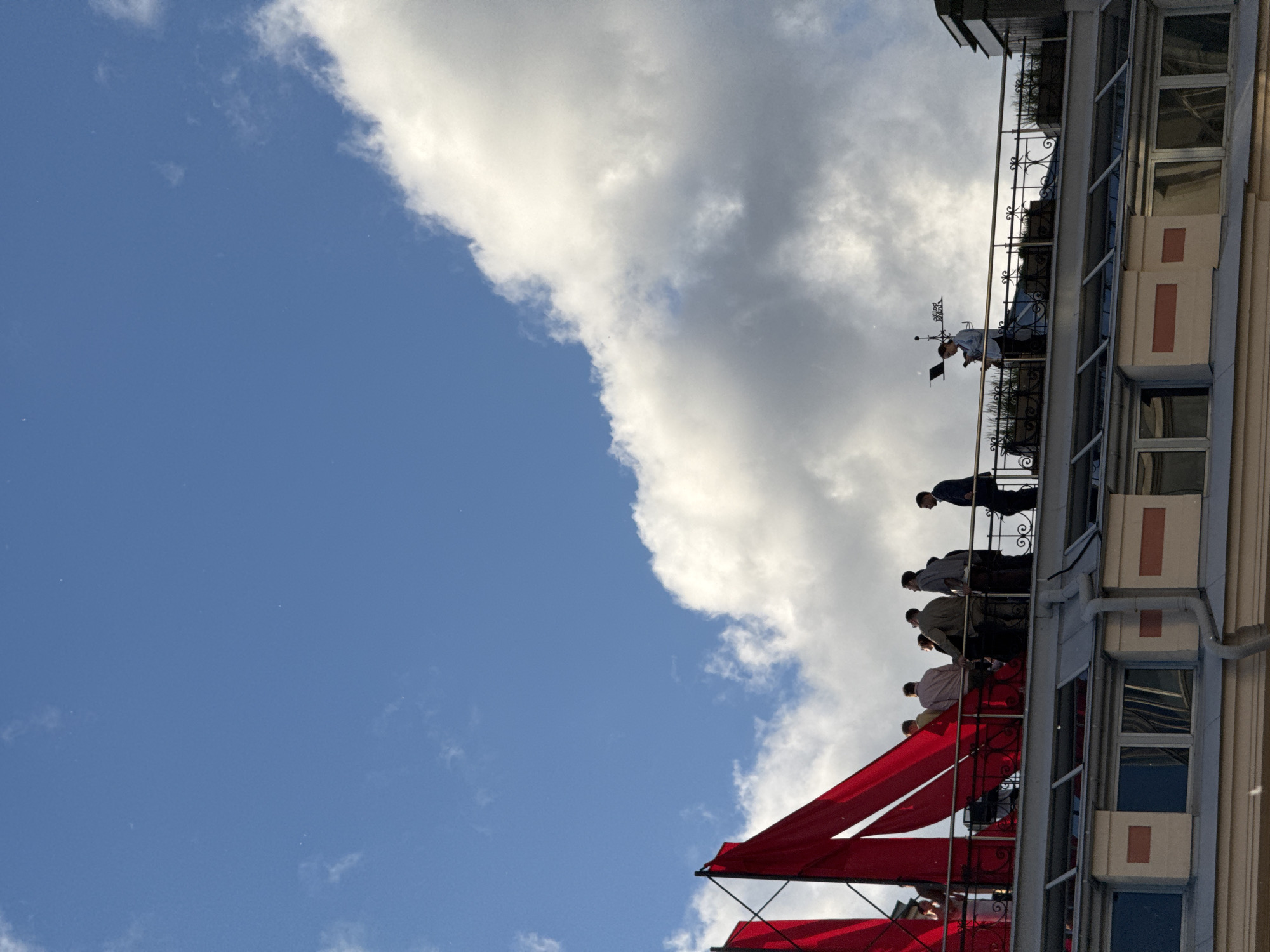 A group of people are gathered on a rooftop terrace with red sails in the background under a blue sky with clouds. The people are standing near a railing, some facing the camera and others looking away. One person on the right side of the terrace is holding a phone. The rooftop has planters with greenery and a weather vane. Below the terrace is a building with large windows.