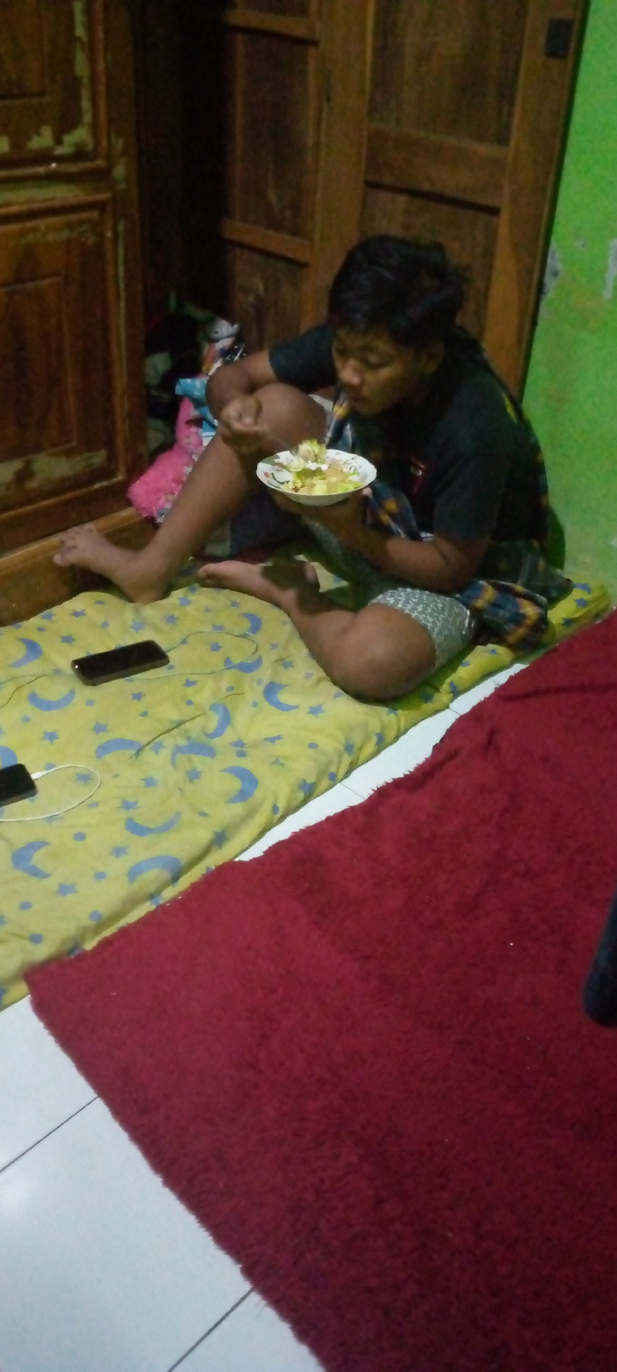 A young person is seated on a yellow blanket with moon and star patterns, holding a plate of food and eating. They are wearing a black t-shirt and patterned shorts. To their left, another blanket, this one red and fuzzy, is spread out. Two black smartphones are visible on the yellow blanket, one with a charging cable connected. Behind the person is a wooden cabinet with its doors open, revealing stacked items. The environment appears to be an indoor space in Sumberwono, Indonesia, likely a bedroom, with a green wall on the right and tiled flooring.