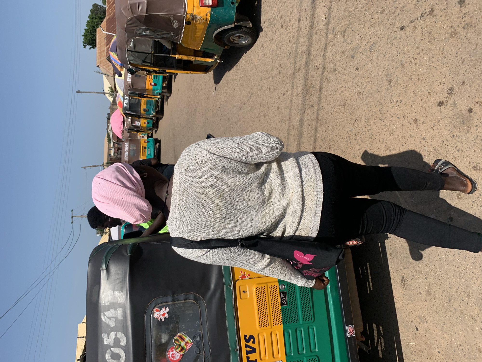 In Gura Top, Nigeria, a woman wearing a light gray sweater, black pants, and a pink headscarf walks away from the camera down a dirt road. She carries a black bag with a pink floral design over her left shoulder. To her left is a green and yellow tuk-tuk with the number "0543" visible on its black roof. A man in a green shirt is partially visible in the tuk-tuk's driver's seat. Several other colorful tuk-tuks are parked in a line on the road ahead of her, some with umbrellas deployed on top. The scene is brightly lit by sunlight, casting shadows on the ground. In the background, buildings with corrugated metal roofs and utility poles are visible under a clear blue sky.