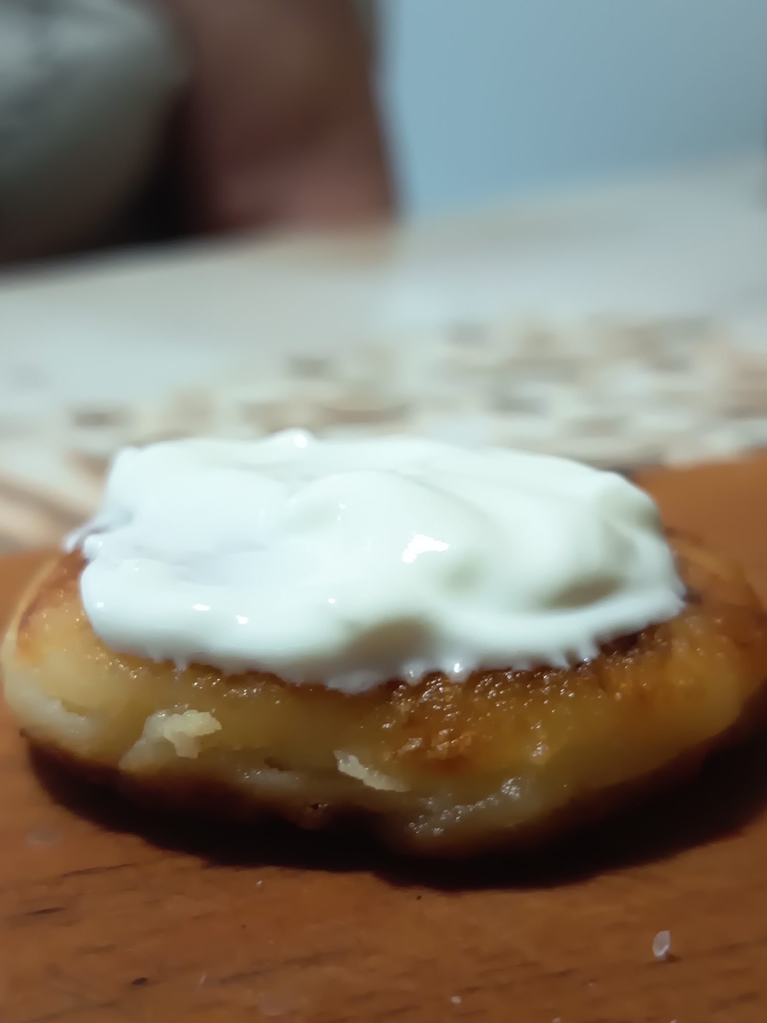 The image displays a close-up view of a single, golden-brown pancake topped with a dollop of white cream. The pancake has a slightly irregular shape and appears to be cooked to a crisp on the edges. The cream is thick and creamy, with visible swirls and indentations from its application. The pancake rests on a wooden surface, likely a table, which is a warm brown color. In the blurred background, a light-colored, textured surface, possibly a tablecloth or placemat, is visible. The overall lighting suggests an indoor setting. The image does not contain any information that would allow for the determination of its location in the city of Izmail, Ukraine.