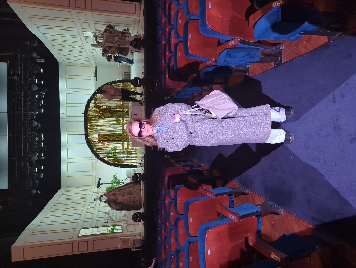 The image depicts the interior of a theater in Moscow, Russia, encompassing an auditorium and a stage. In the foreground, a blonde-haired woman with dark sunglasses stands centered in a dark blue carpeted aisle. She wears a grey tweed-like long coat over a blue patterned shirt, light-wash jeans, and dark shoes, holding a light-colored tote bag with her arms crossed. Rows of unoccupied seats, featuring red wooden backs and blue upholstered cushions, line both sides of the aisle.
The stage in the background is set with white paneled walls accented by gold trim. On the left side of the stage, a large, dark, draped figure or sculpture is positioned next to a white chair and greenery. Centrally, a large semi-circular archway decorated with vertical gold tinsel streamers forms a backdrop, with greenery and a microphone stand placed in front. Another draped figure or prop is visible on the right side of the stage. Multiple stage lights are present on the floor and suspended overhead. Two individuals are on stage: one person in a light-colored top stands near the central arch holding a guitar, and another individual is partially visible towards the right, suggesting a rehearsal or setup phase.