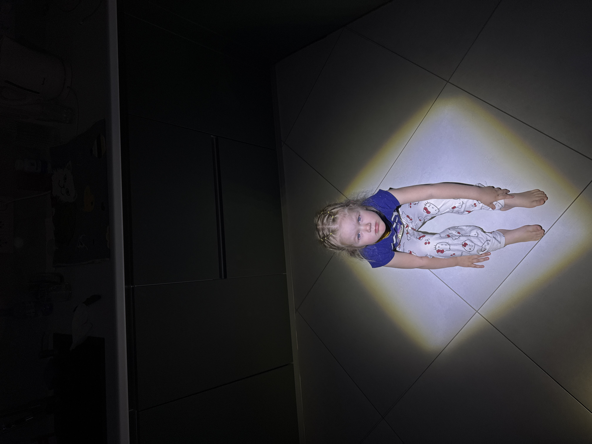 A fair-skinned child with blonde braids sits on a tile floor under a bright, square-shaped light. The child wears a blue t-shirt and white pants with a repeating Hello Kitty pattern. The setting appears to be a room with dark cabinets on the left side. The location may be Ферма, Russia.
