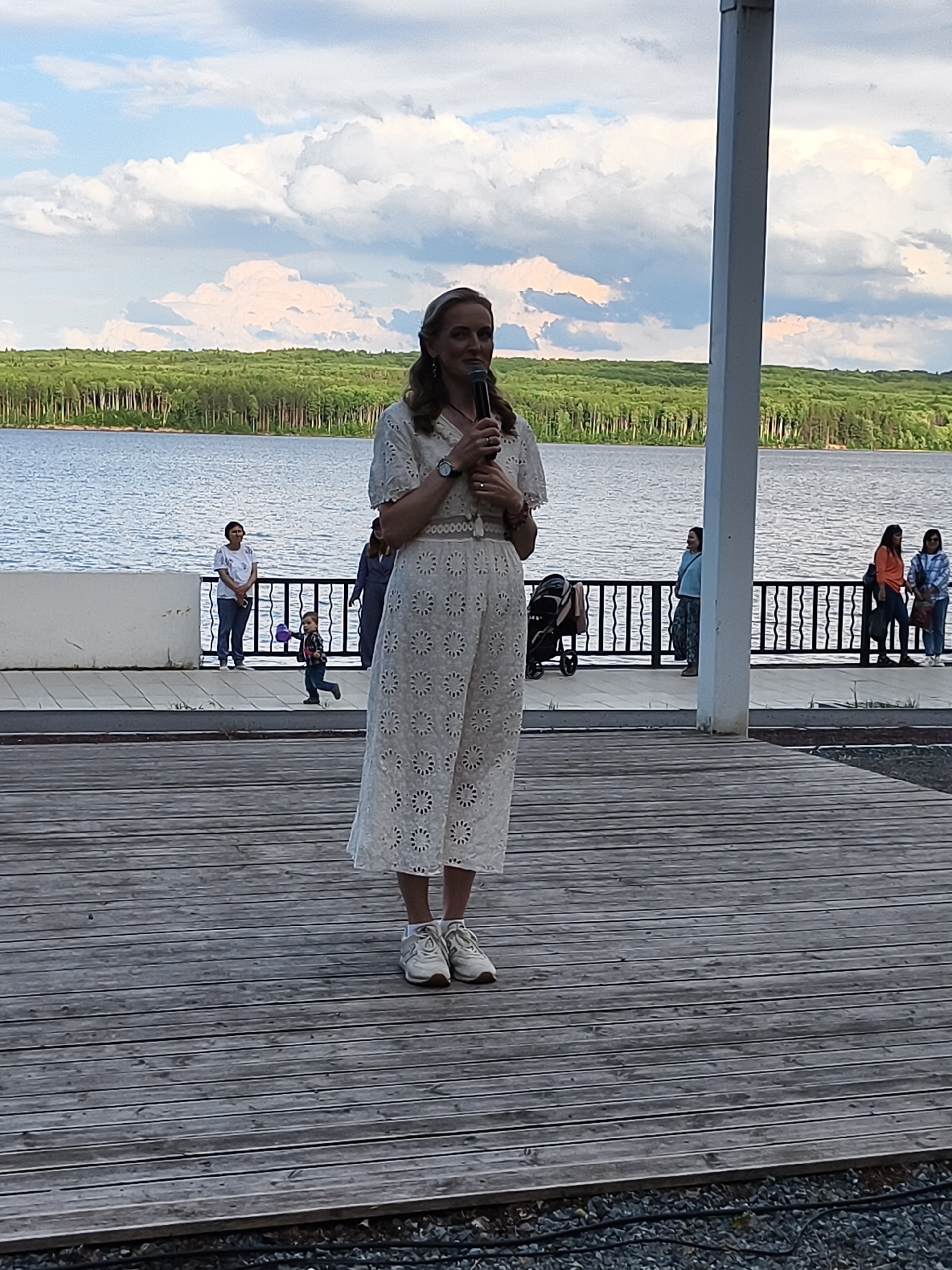 A woman in a white dress and white sneakers is standing on a wooden deck, holding a microphone. She is looking to her right and appears to be speaking. Behind her, a lake is visible, with a line of trees on the far shore. Several other people are standing near a railing along the edge of the deck, some of whom are looking towards the woman. The sky is cloudy. The scene is likely outdoors, possibly in a park or recreational area in Chaikovsky, Russia, given the presence of a lake and natural scenery.