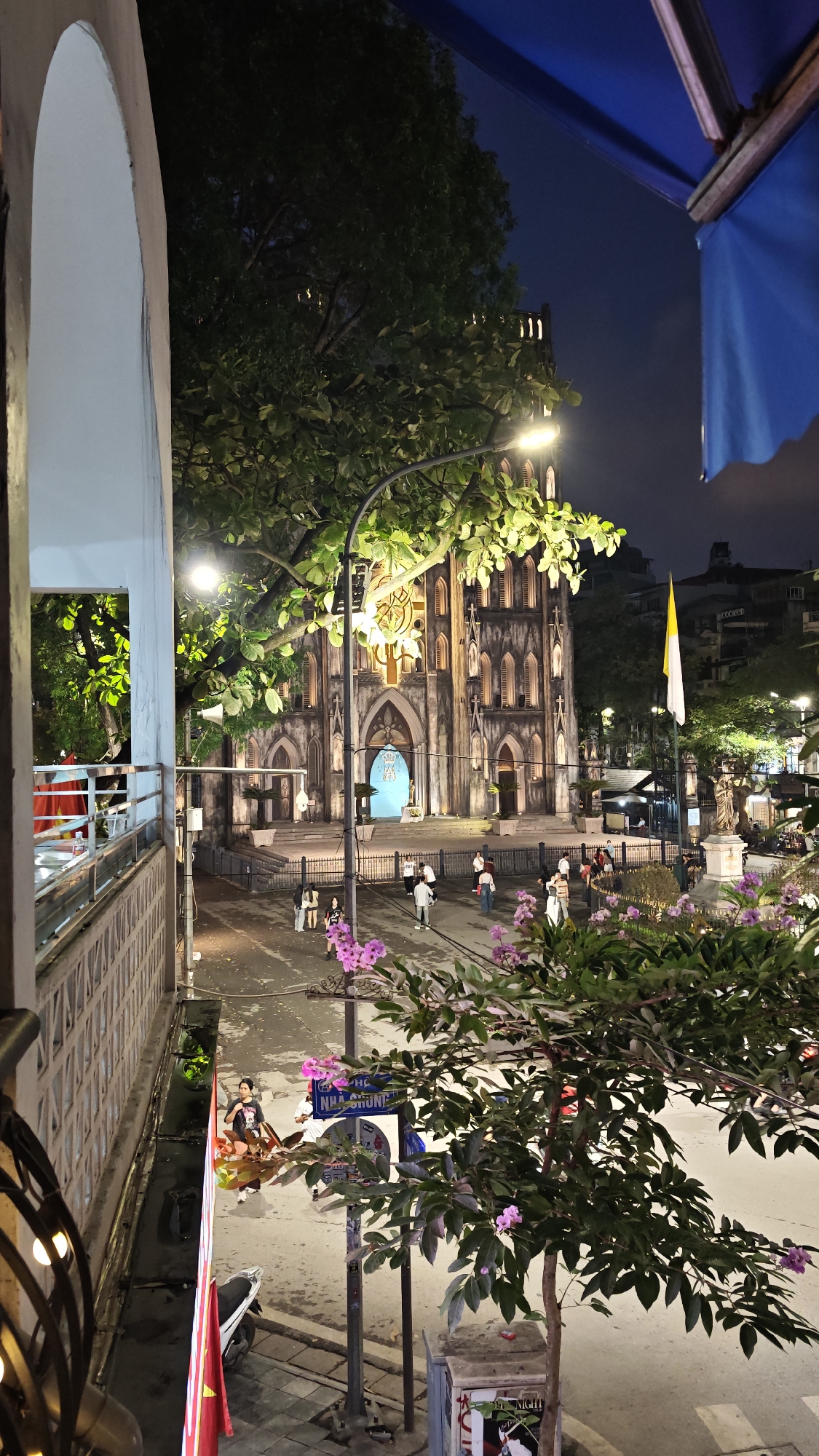 Captured at night in Hà Nội, Vietnam, a Gothic-style cathedral dominates the scene, illuminated with spotlights. Pedestrians are seen on the sidewalks and in the street. The camera is positioned on a balcony or elevated platform with a partial view obstructed by foliage. Street signs are visible, and a Vietnamese flag is displayed to the left of the frame. A street lamp stands centrally, and a blue canopy is at the top right corner. The environment suggests a lively urban setting, with the church as a focal point.