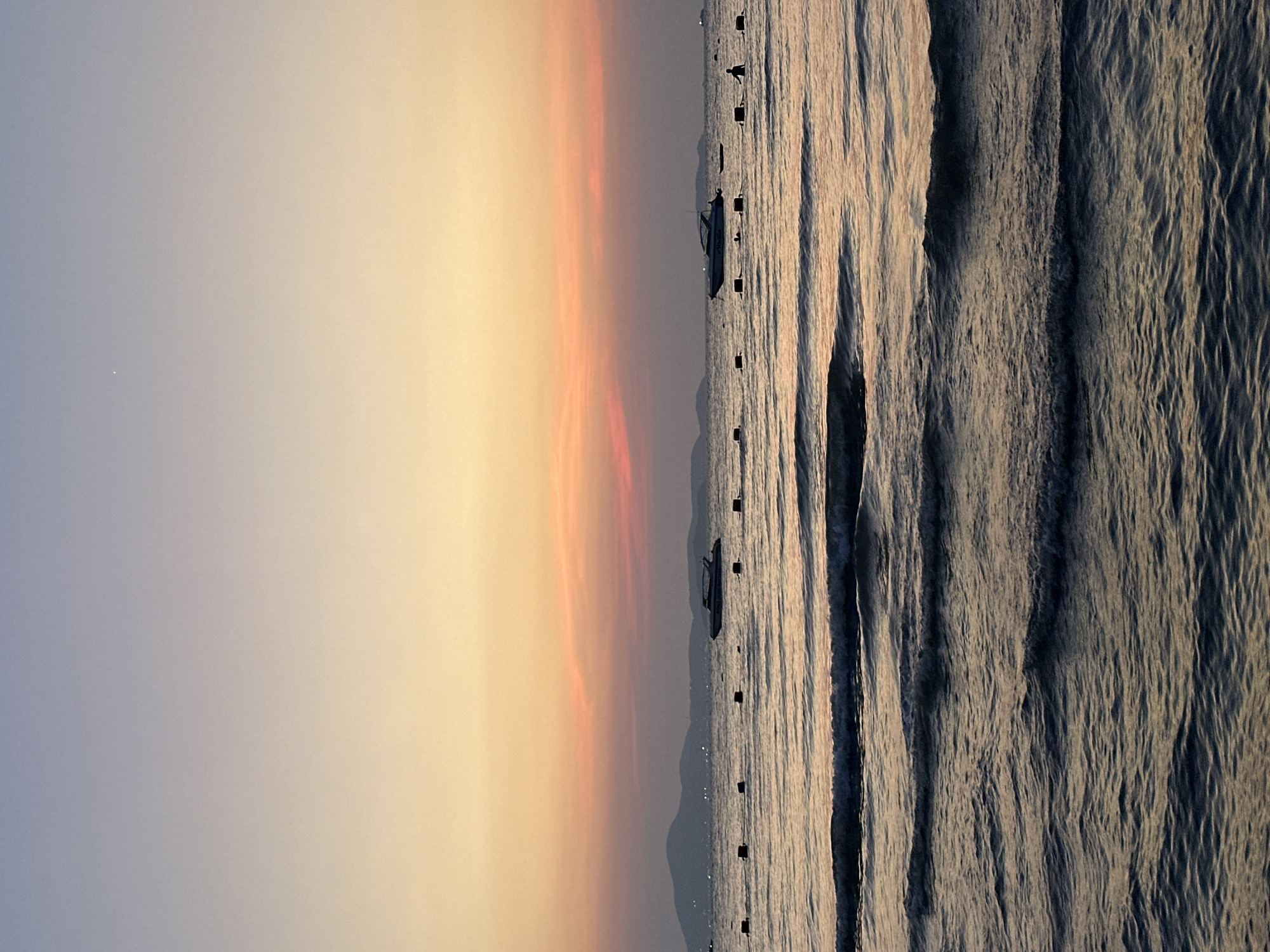 The image depicts a sunset over the ocean in Pattaya City, Thailand. The sky transitions from light blue to golden yellow and pink hues. Several boats are visible on the water, delineated by dark buoys marking a perimeter. The ocean surface is textured with waves reflecting the fading sunlight. Silhouetted mountains are present on the horizon.