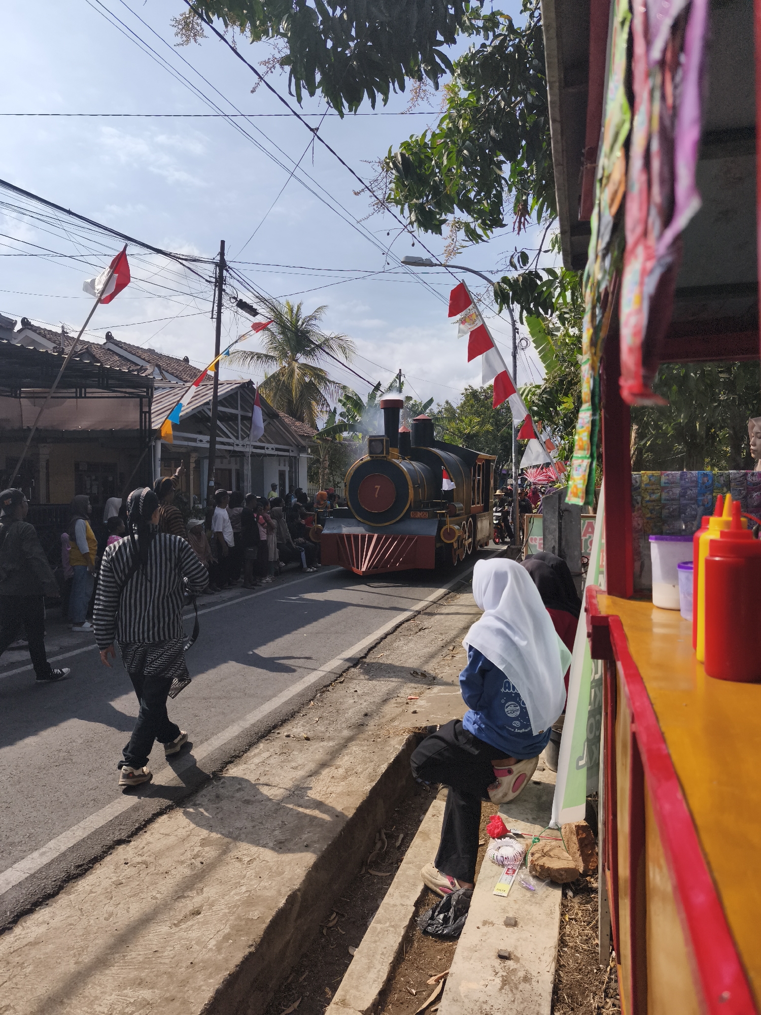 This is a street-level view of a parade in Kutoarjo, Indonesia, featuring a decorative steam locomotive. The locomotive, painted black and red with gold accents, displays the number "7" on its front. It is adorned with Indonesian flags, red and white banners, and is emitting steam. The parade is taking place on a paved road, with houses and trees lining the sides. A crowd of people, dressed in casual attire and some wearing traditional clothing like the woman in the foreground with a striped shirt and headscarf, are gathered along the street to watch. Several Indonesian flags and colorful pennants are strung across the road and on utility poles. To the right of the frame, a food stall or vendor cart is visible, with colorful packets of items hanging from it and condiments like red and yellow squeeze bottles on the counter. A young woman wearing a blue sweater and a white hijab is seated on the curb, facing away from the camera. The sky is blue with scattered clouds.
