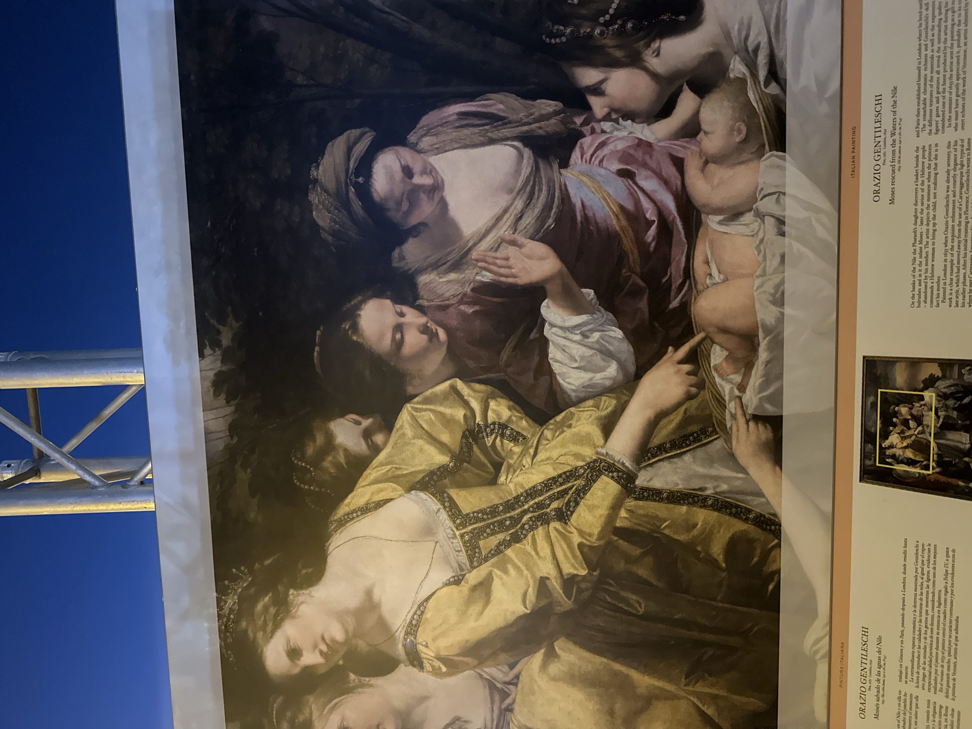 The image displays a section of a large painting, framed by a metal structure and a dark blue sky, suggesting it is outdoors. The painting depicts a biblical scene with several women gathered around a baby. The central focus is a baby lying on its back, wrapped in white cloth. To the left, a woman in a shimmering gold dress, adorned with dark, intricate trim and pearls, gazes down at the baby, with her left hand pointing towards it. Behind her, another woman in a mauve dress with a draped neckline also looks at the baby. To the right, two other women are partially visible. One in a dark head covering leans in close, her face obscured, while another woman with dark hair and a bejeweled headdress is seen in profile. The background of the painting is dark and appears to be a landscape with foliage. Below the painting is a display board with text, including titles like "PINTURA ITALIANA" and "ORAZIO GENTILESCHI," and descriptions of the artwork. The text mentions "Mazarrón, Spain," suggesting the painting is displayed or exhibited in this city.