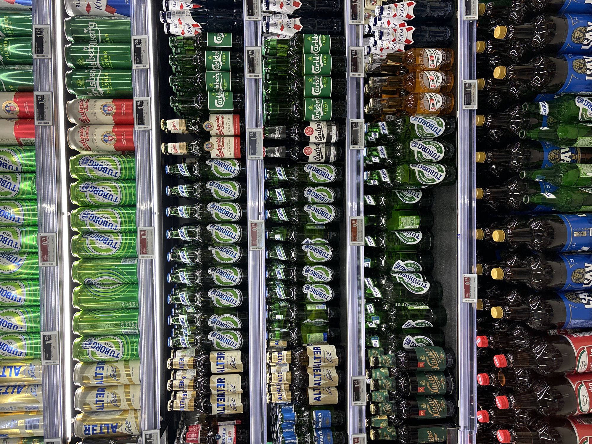 The image presents a well-stocked refrigerator display in a retail setting, likely a supermarket or convenience store. The shelves are filled with various brands and types of beer, arranged in neat rows. Prominent brands include Tuborg, Carlsberg, Budweiser Budvar, and Alter. Price tags are visible next to some of the items. The beers come in both cans and bottles.
While the image provides details about the beer selection and pricing, it doesn't contain information about people, specific actions, interactions, or any clues that would help pinpoint a location in Budva, Montenegro. Therefore, it's impossible to confirm the location of the image based on visual cues.